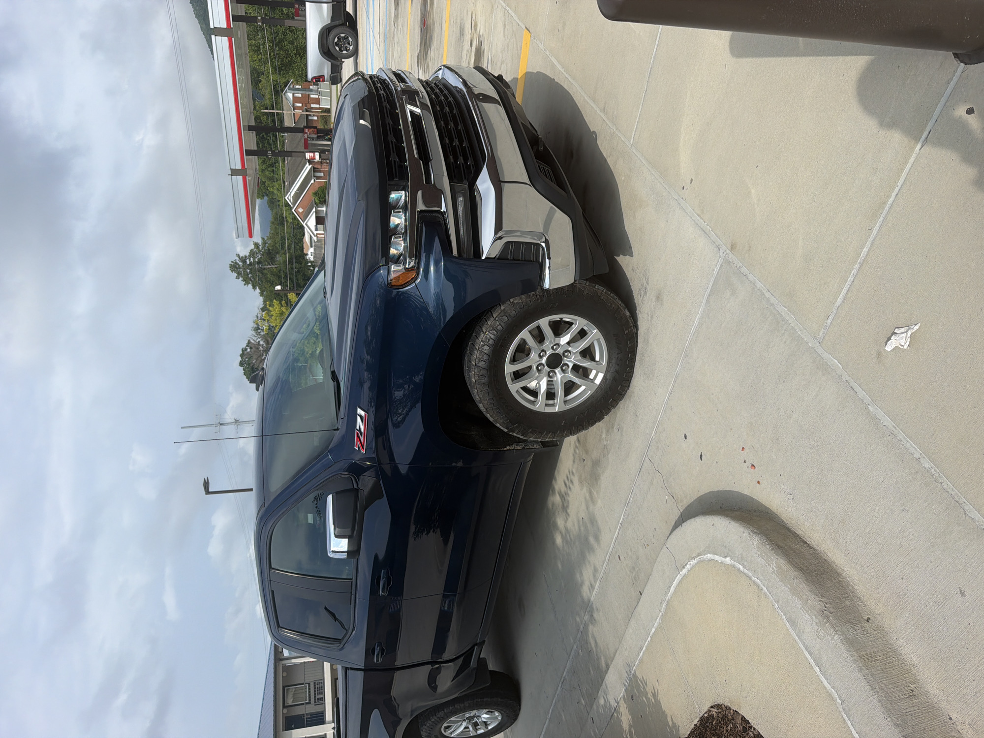 A dark blue Chevrolet Silverado pickup truck is parked in a concrete lot. The truck is facing towards the right side of the frame. The truck has chrome accents on the grille, bumper, and side mirrors. The Z71 badging is visible on the front fender. In the background, a gas station with a red and white canopy is visible, along with some trees and buildings. The sky is cloudy. The scene is consistent with a gas station parking lot in Chattanooga, Tennessee, United States of America.