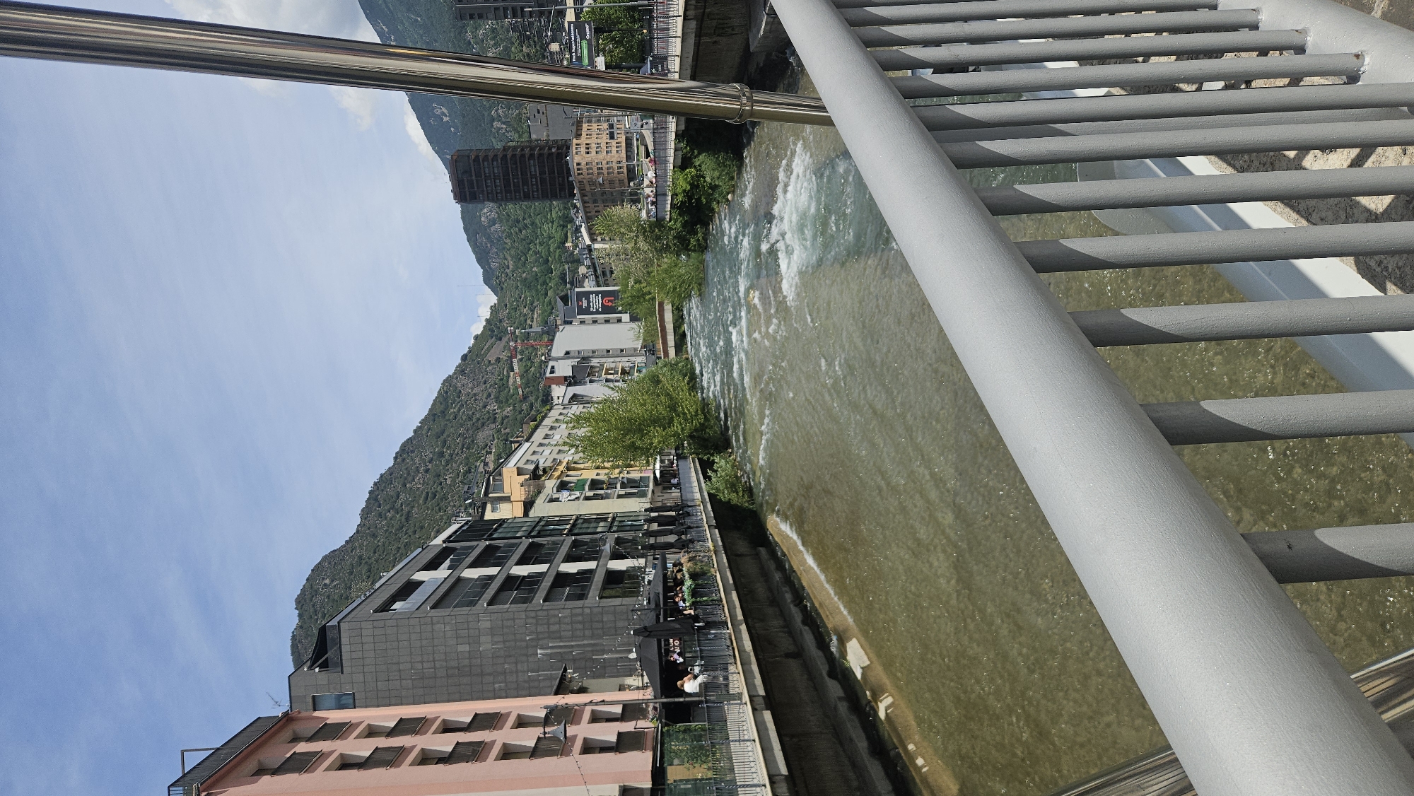 The image presents a detailed view of the Gran Valira river flowing through Andorra la Vella, Andorra, observed from a bridge on a clear, sunny day. In the immediate foreground, a grey metal bridge railing, composed of a horizontal top bar and numerous vertical balusters, extends from the lower right across the bottom of the frame. To the railing's left, a reflective silver-colored vertical support column is partially visible.
The midground is dominated by the river, characterized by its murky, brownish-green water and numerous areas of turbulent white rapids, indicative of a strong current. The left bank is densely urbanized, featuring a promenade lined with buildings. The closest structure on the left is a multi-story light pink building with balconies. Adjacent to it stands a modern, dark grey building with extensive fenestration and balconies. Further along the bank, a series of diverse commercial and residential buildings of varying colors and styles are visible. Below these structures, an outdoor cafe area with multiple black umbrellas and seated patrons is active, and several individuals are seen walking along the promenade.
The right bank, extending into the distance, also features urban development, including a tall, dark brown building prominent against the mountainous backdrop. Green foliage and trees line sections of both river banks. Two bridges span the river in the distance: a smaller, light-colored bridge closer to the center, and a wider bridge further downstream. A red construction crane is discernible high on the mountain slope on the right side of the image. The background consists of steep, verdant mountains, covered in trees and exposed rock, rising directly behind the city. The sky above is largely clear and blue, with scattered white clouds.