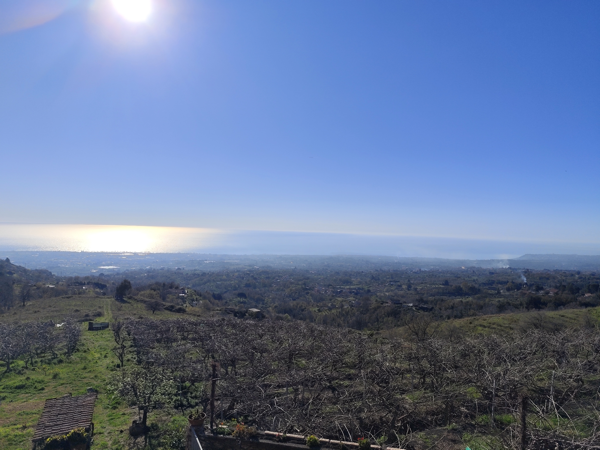 A daytime, long-shot aerial view of a landscape in Giarre, Italy, features an extensive view of a valley with a city in the background and a bright blue sky. A body of water that is likely the sea, reflects intense sunlight on the horizon, creating a shimmering effect. In the foreground, there's a mix of terrain with patches of green grass, bare trees, and perhaps a plantation or an orchard in a state of winter dormancy. A structure with a red-tiled roof is partially visible in the lower left corner. The city visible in the valley appears dense, with a muted color palette due to the distance and possibly atmospheric haze. The sky transitions from light blue to a deeper blue, and the sun's glare dominates the upper part of the frame.
