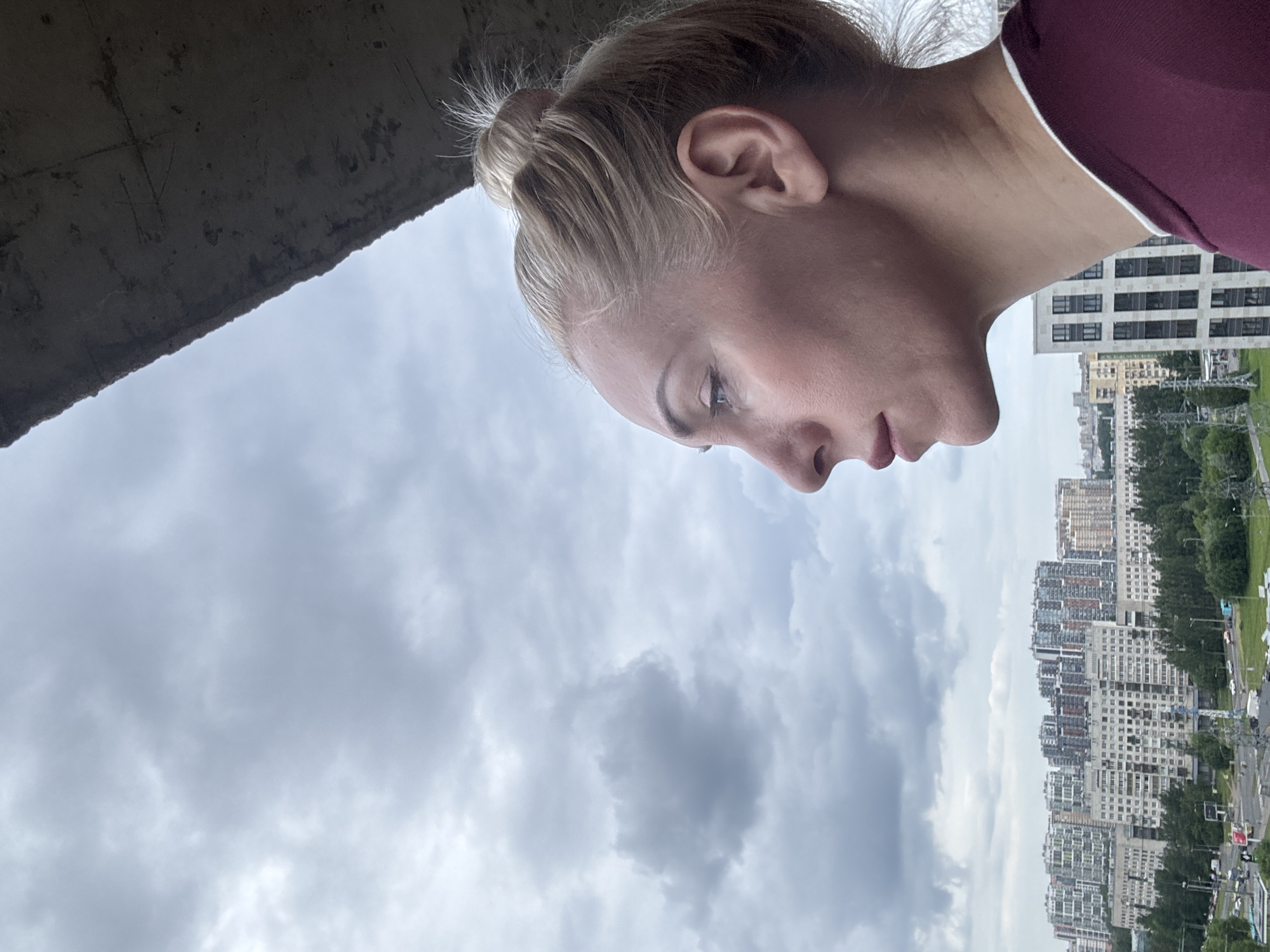 A blonde woman with her hair in a bun looks to the left against a cloudy sky. She is in profile and wears a maroon shirt with a white collar. Below her is a cityscape with numerous apartment buildings and trees. A concrete overhang is visible in the upper right corner of the image.