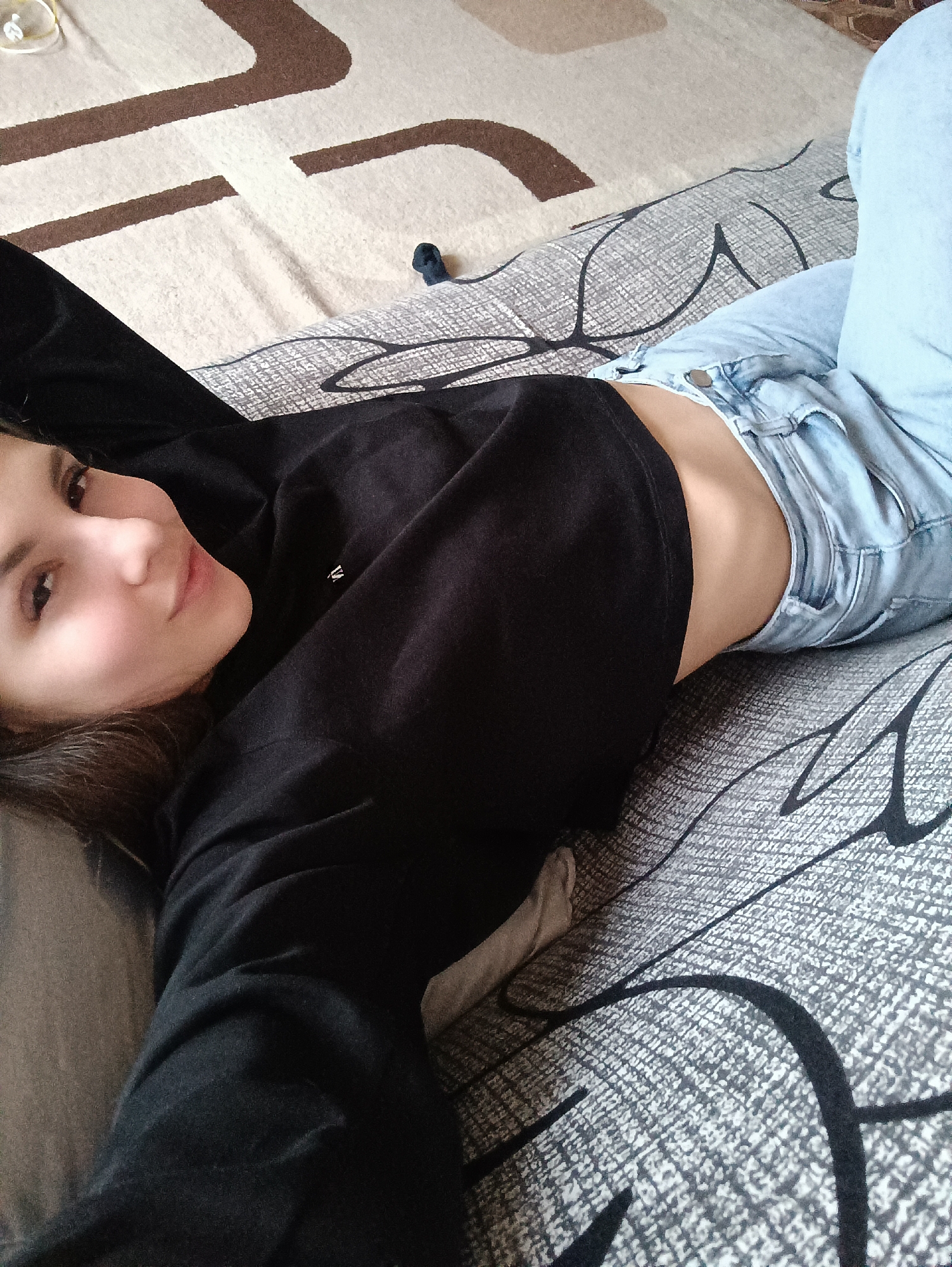 A young woman is lying on a bed, positioned at an angle with her head towards the left of the frame. She is wearing a black cropped sweatshirt and light blue jeans. The bed has a patterned gray and white comforter. A portion of a tan and brown rug is visible in the upper left corner. The location appears to be an indoor residential setting, possibly in Chaikovsky, Russia, based on the provided information.