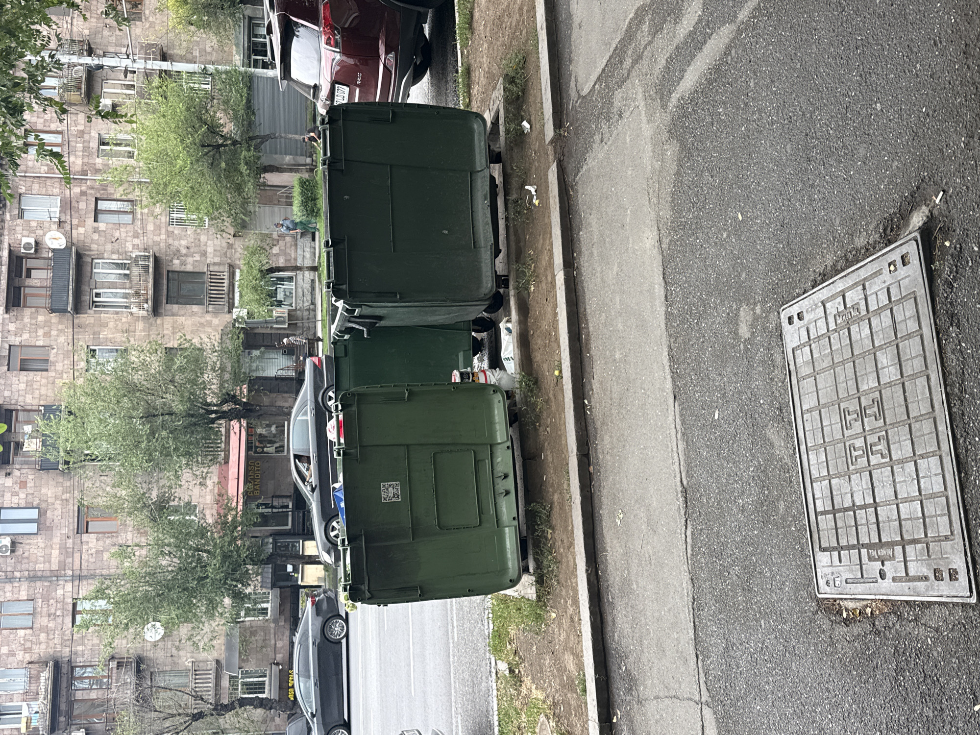 This image depicts a street scene in Yerevan, Armenia, characterized by a row of apartment buildings in the background. In the foreground, several large, dark green dumpsters are positioned on a patch of dirt and grass alongside a sidewalk. These dumpsters are partially filled and show signs of use. To the left of the dumpsters, a street with parked and moving cars is visible, indicating an urban environment. A manhole cover with a grid pattern is situated on the asphalt in the lower part of the frame. The overall scene suggests a typical urban setting with residential buildings, public services like waste disposal, and daily street activity.