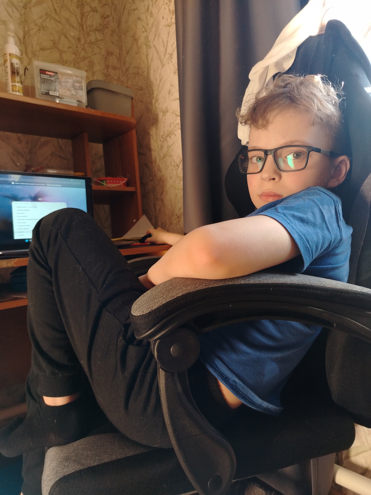 A young boy with light skin, short light-brown hair, and glasses is seated in a gray and black office chair, facing toward the camera and slightly angled to his right. He is wearing a blue t-shirt and black pants. His left leg is propped up on the chair, and his right arm is resting on the chair's armrest. Behind the boy, there is a desk with a laptop on it. The wall behind the desk is covered with patterned wallpaper. A shelf above the desk holds various containers and objects. A dark gray curtain is visible to the right of the boy. The location in the city of Nizhnyaya Gar', Russia cannot be determined from this image.