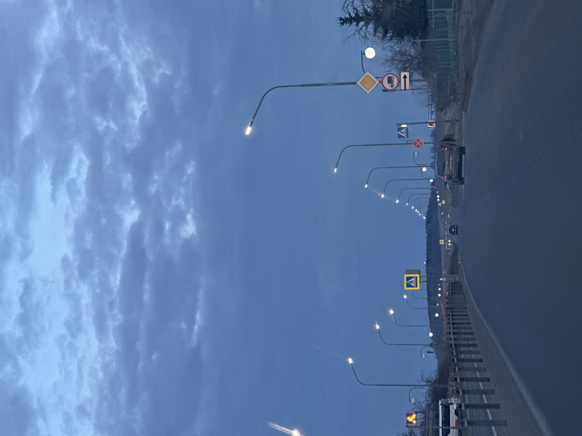 The image depicts a multi-lane asphalt road at twilight or dawn in Liozna, Belarus, viewed from a slightly elevated position, looking straight ahead down the road. The sky above is dark blue, filled with numerous, diffused grey clouds indicating overcast conditions.

The road, appearing dark grey, extends into the distance, lined on both sides by illuminated streetlights with bright LED fixtures, creating a receding tunnel of light. A metal guardrail separates the left side of the road from a shoulder, where a white bus is partially visible.

On the right side of the road, a dark grey sport utility vehicle (SUV) is moving away from the viewer in the rightmost visible lane. Two indistinct human figures are walking on the paved shoulder immediately to the right of the SUV.

Various road signs are present: a prominent yellow diamond-shaped "main road" sign, a red circular "no trucks" sign, and a white rectangular arrow sign pointing right are mounted on a pole on the right. Below these, a rectangular sign states "ПЛАТНАЯ ДОРОГА" and "TOLL ROAD" in white text on a blue background, indicating a toll route. Multiple yellow square signs with black pedestrian symbols (pedestrian crossings) are visible on both sides of the road. A red circular prohibitory sign is also visible further down on the right.

Bare-branched trees and a dark coniferous tree stand along the right side of the road. The distant horizon shows a gentle rise in elevation, with distant lights marking the path of the road.