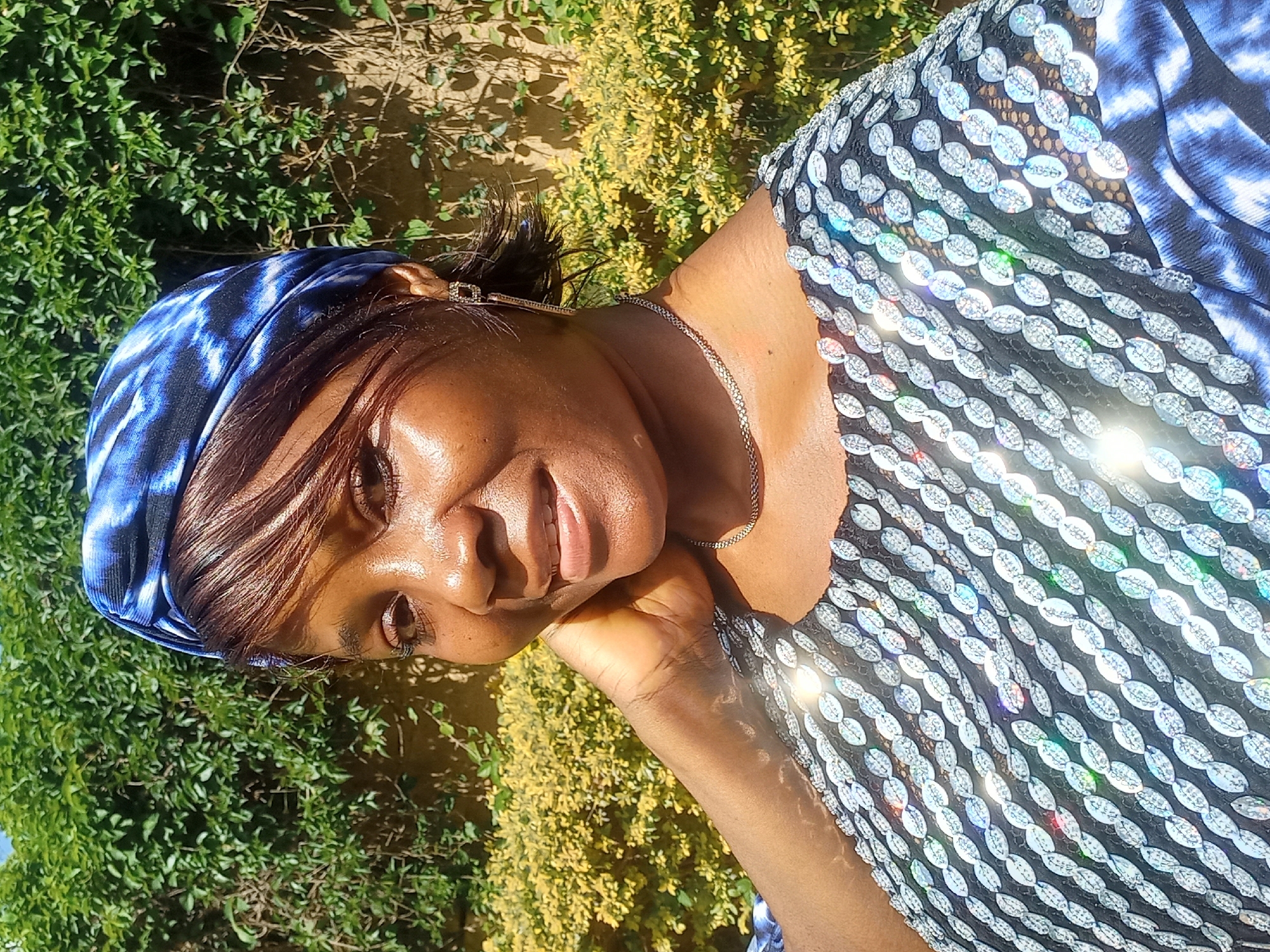 A medium shot, taken outdoors in daylight, features a Black woman from the chest up, posing for a selfie. She is wearing a blue and white tie-dye headwrap and a black top adorned with rows of silver sequined embellishments. The background consists of a dense green hedge and a wall. Her right hand is placed behind her neck, and she is looking towards the camera with a slight smile. The overall impression is one of warmth and natural light, suggesting a pleasant outdoor setting, possibly in the city of Jos, Nigeria, given its typical climate and vegetation.
