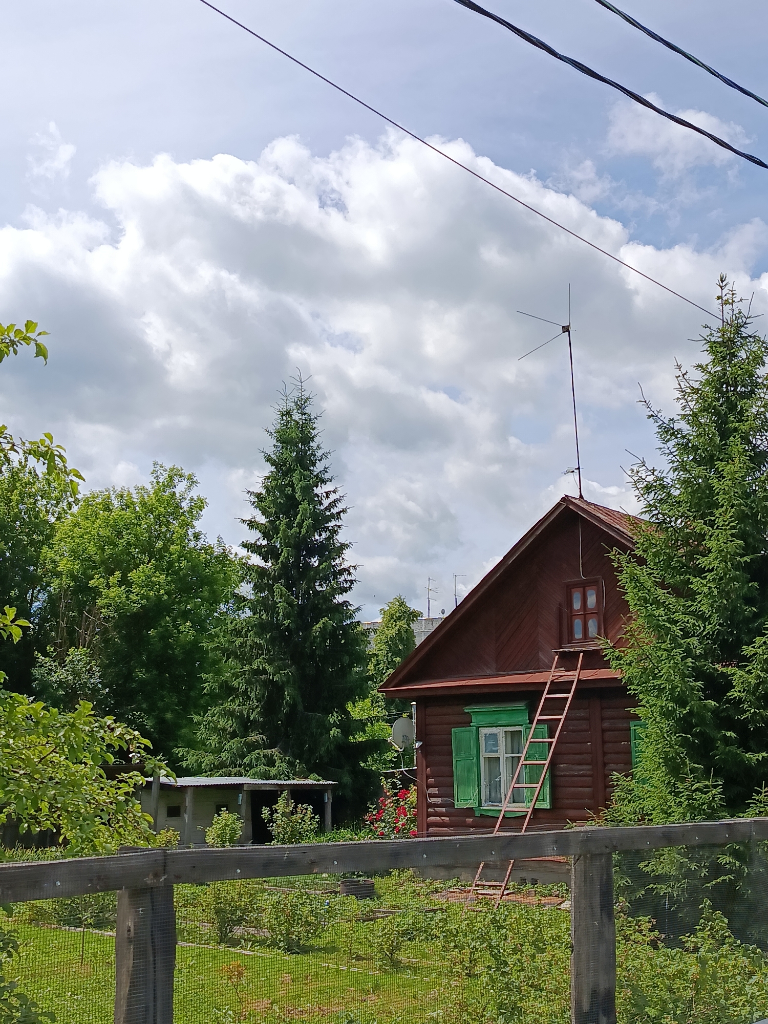 The image shows a wooden house in Vereya, Russia, with a brown exterior and green shutters on one of its windows. A ladder leans against the house, reaching towards an attic window. The house is surrounded by lush green trees and a garden enclosed by a wooden fence. The sky is partly cloudy, with white clouds scattered across a pale blue background. Power lines are visible in the upper part of the frame, running across the sky. In the background, a small shed with a metal roof can be seen. An antenna is mounted on the roof of the house. The overall scene depicts a rural or suburban setting in Vereya, Russia, with a focus on the house and its natural surroundings.