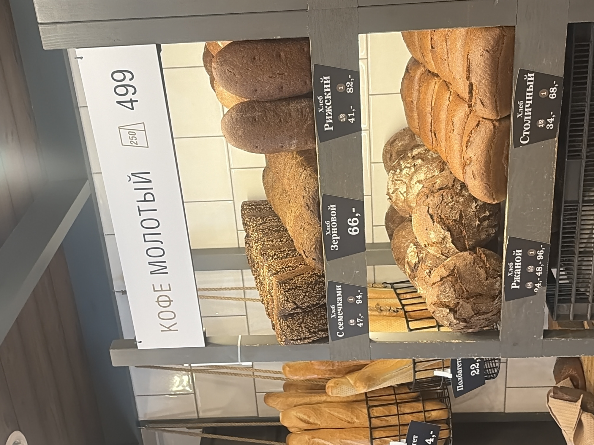 The image shows a bakery shelf in Saint Petersburg, Russia. A sign on the shelf reads "КОФЕ МОЛОТЫЙ" (ground coffee) and lists the price as 499. The bakery offers a variety of bread including: bread with seeds, Grain bread, Riga bread, Rye bread, and Metropolitan bread. Each bread type has the price of a half and whole loaf listed in rubles. There are loaves and baguettes on the shelves.