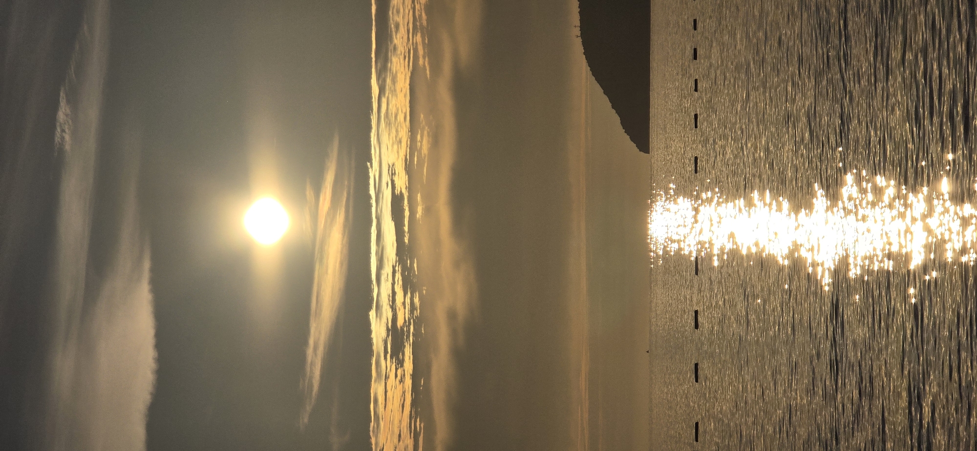 The image depicts a coastal sunrise or sunset scene in Bar, Montenegro, characterized by warm, golden light across the sky and sea. The upper portion of the frame features a bright, intense, light-yellow sun, appearing somewhat square due to glare, positioned in the upper center-left against a sky that transitions from dark grey-blue at the top to golden-orange hues closer to the horizon. Horizontal bands of illuminated, streaky, and layered clouds are visible, particularly in the mid-sky. Below these, a hazy, light brown-orange atmosphere extends to the distant horizon.
The lower half of the image shows a body of water, likely the Adriatic Sea, with gentle ripples. A prominent, vertical path of sparkling, bright yellow sunlight reflects on the water's surface, extending from near the horizon towards the bottom edge of the frame. A line of approximately seven to eight dark, rectangular buoys or markers stretches horizontally across the mid-ground of the water, defining a boundary. A small, dark, indistinct object, possibly a distant boat or buoy, is visible further left on the water.
On the right side of the image, a dark, silhouetted landmass, appearing as a hill or small mountain, slopes down to meet the water. Two faint, pole-like structures are discernible near its peak against the lighter sky. No individuals or animals are visible.