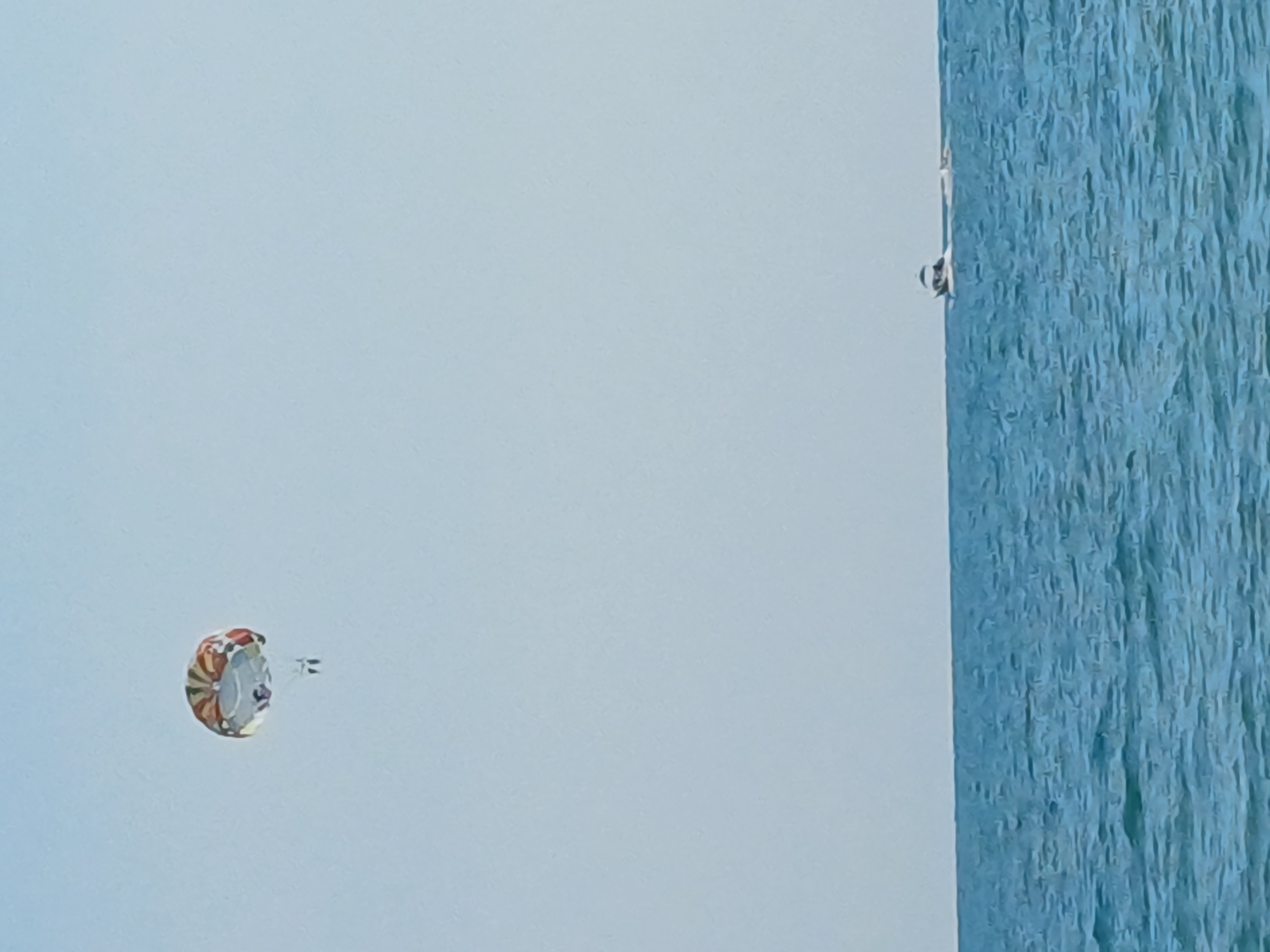This media file captures a parasailing activity in Sochi, Russia. The scene is dominated by a light blue sky, with a section on the right displaying a textured, vertical blue surface, possibly a building or a water feature. In the sky, a colorful parasail with red, white, and yellow sections is visible, trailing behind a small boat or jet ski which is also in motion, indicated by the faint white lines behind it. A person is seated in the harness beneath the parasail, enjoying the flight. On the far right edge of the image, near the blue textured surface, a small boat with a person on board is also visible, suggesting it's the towing vessel. The overall impression is one of a sunny day, ideal for water sports. There is no visible text in the image.