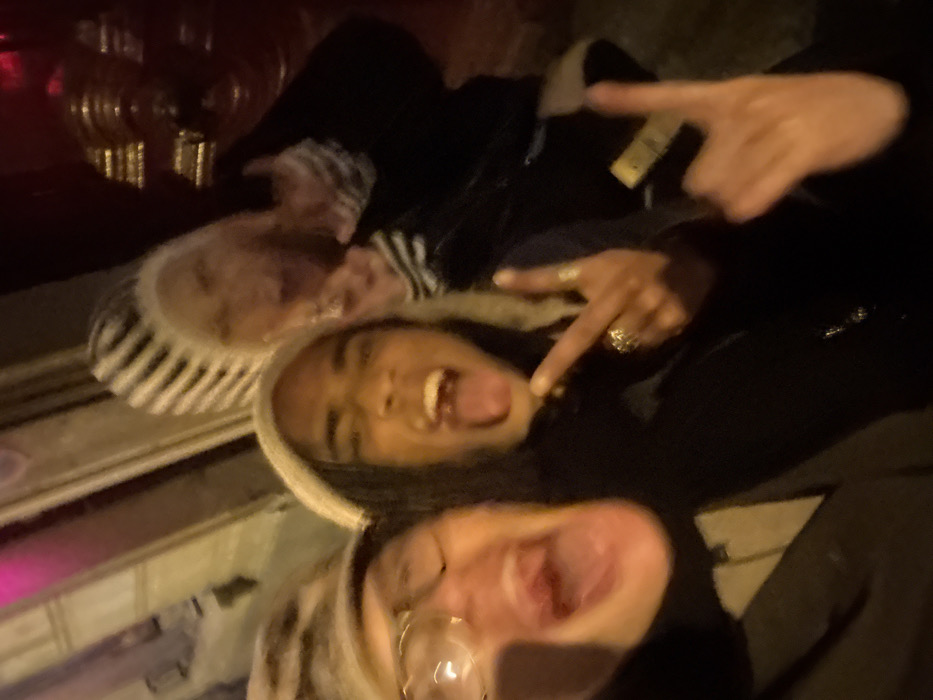 Three people pose for a selfie. The person in the middle has brown hair and is wearing a white hooded sweatshirt. They are sticking their tongue out and pointing at the camera. The person on top is wearing a black hooded jacket and has a ring on their finger. The person on the bottom is wearing glasses and also has their mouth open. All three people are smiling. The picture is blurry and dark.