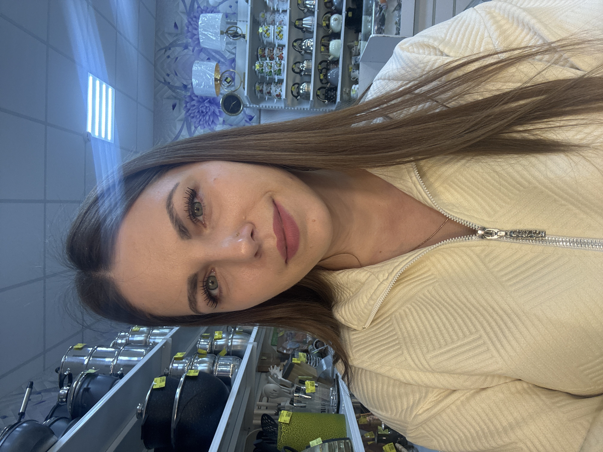 A woman is taking a selfie inside a store. Shelves displaying cookware and kitchenware are visible behind her, along with other household goods on higher shelves. The location appears to be Klin, Russia. She is wearing a light-colored textured jacket.