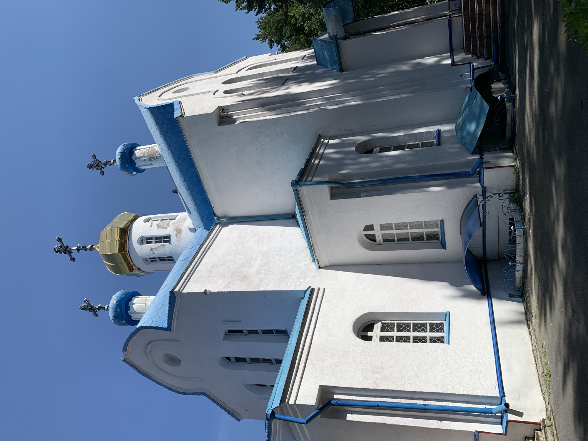 A white church with a blue roof and gold domes stands under a clear blue sky. The church has several arched windows and is decorated with blue trim. A few trees are visible in the background. The image was taken in the city of Prokopyevsk, Russia.
