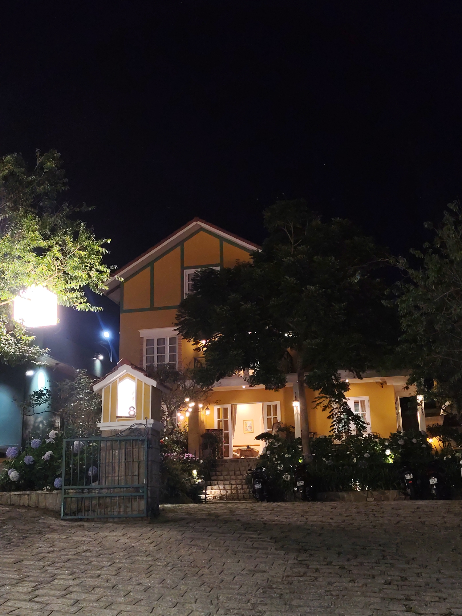 The image captures a two-story building in Unknown, Unknown, at night. The building is painted in a yellow hue with white trim around the windows and doors. A brown roof complements the color scheme. A decorative, small structure that is illuminated stands in front of the building. To the left, hydrangea bushes are in bloom. A metal gate guards the property entrance. The street in front of the building is cobbled. Trees flank the building, their foliage obscuring parts of the facade. Additional motorcycles are parked behind a shrubbery hedge in the foreground.
