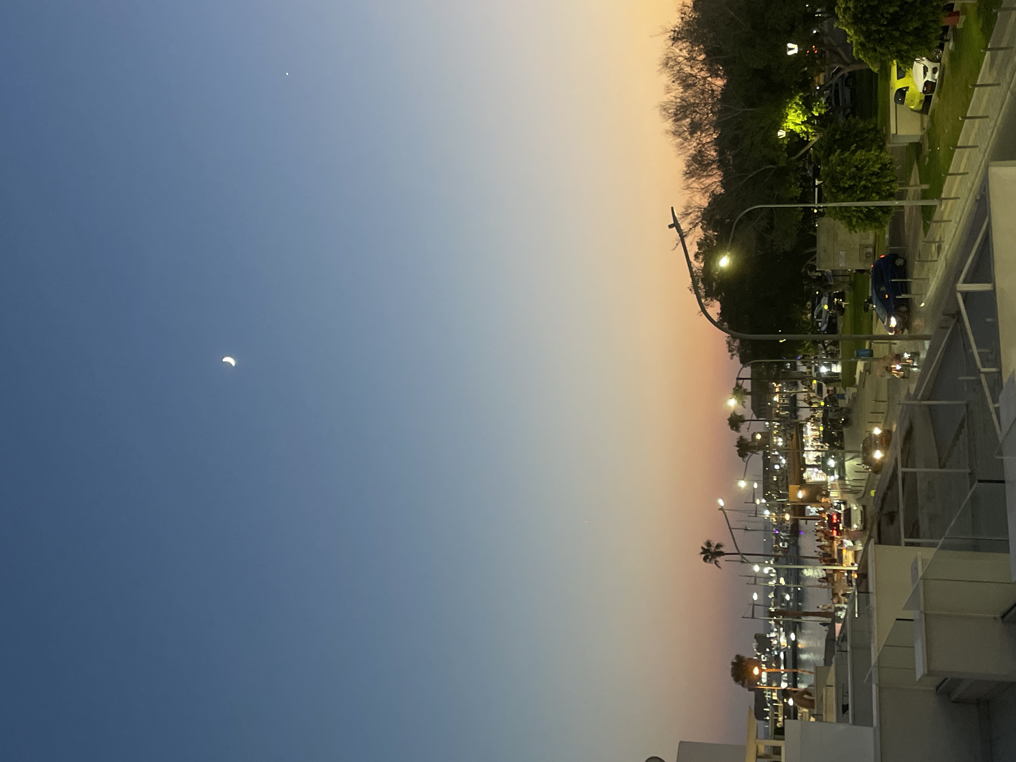 This image captures a coastal scene in Paphos Municipality, Cyprus, at dusk. The sky displays a gradient of colors, transitioning from a deep blue at the top to orange and pink near the horizon. A crescent moon and a small bright star are visible in the upper sky.
The foreground features the edge of a building with white balconies. Below, a street is lined with cars and streetlights. Several vehicles, including a blue hatchback and a yellow car, are parked or driving. People are scattered along the waterfront area.
In the midground, a harbor is filled with numerous boats and yachts, their lights reflecting on the water. Palm trees are interspersed with buildings along the coastline. The background consists of more trees and structures, suggesting a developed urban area adjacent to the sea. The overall atmosphere is that of a lively evening in a coastal town.