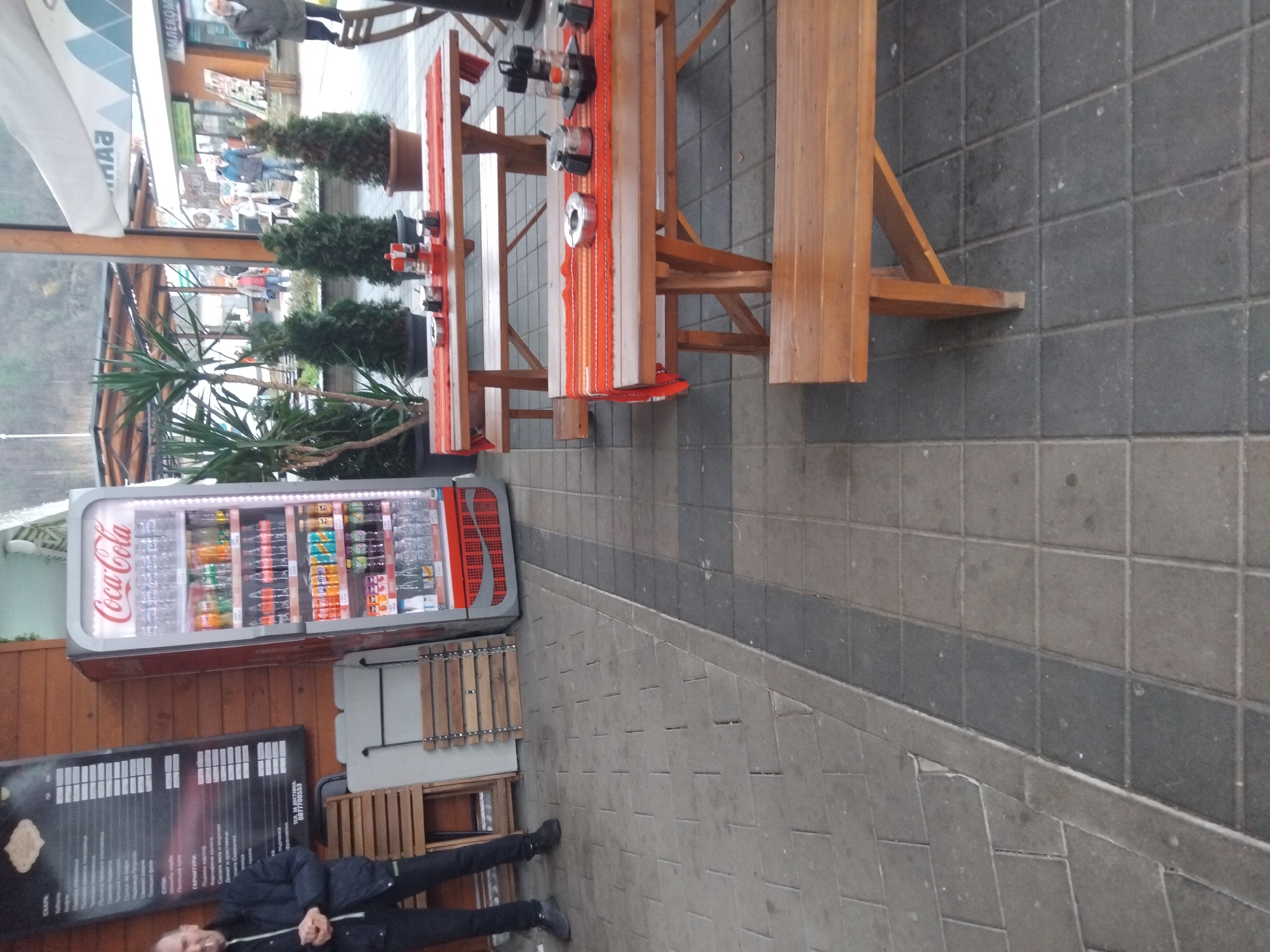 The image presents an outdoor scene in Troyan, Bulgaria, situated on a paved area. A Coca-Cola refrigerator, stocked with beverages, is positioned against a wooden wall, adjacent to a menu board. A man dressed in dark clothing lies near the board. Several wooden picnic tables and benches are arranged on the right side of the image, some adorned with orange-striped runners and various objects. Green plants in pots are visible on the tables and nearby. In the background, there's a glimpse of a street scene with pedestrians and buildings.