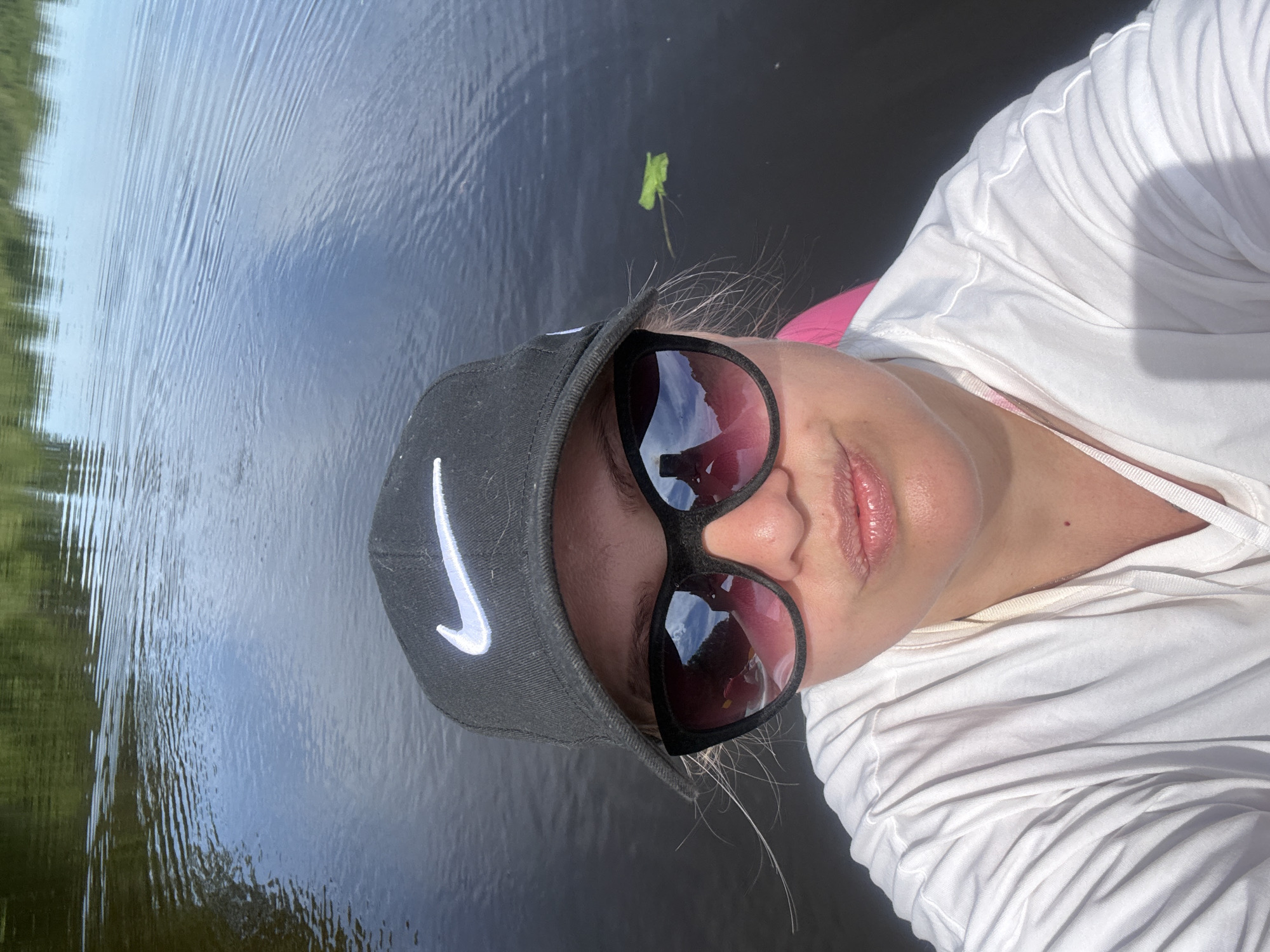 A close-up selfie depicts an individual positioned on a body of water, likely a lake or river. The person wears a black baseball cap featuring a white Nike swoosh logo on the front. Black-framed, pink-tinted sunglasses obscure the upper half of the face, reflecting a bright sky, distant green foliage (possibly hills or trees), and a red/pink object, consistent with the interior of a watercraft, along with the device used to take the photograph. The person's lips are slightly pursed and appear glossy. A white, long-sleeved shirt with visible creases covers the upper torso. Behind the individual's right shoulder, a bright pink surface, possibly part of a kayak or boat, is visible. The surrounding water is dark with distinct ripples, reflecting patches of blue sky and distant green vegetation. A single small, green leaf floats on the water to the right of the person. The lighting is bright and natural, casting shadows on the shirt. The image contains no identifiable landmarks, structures, or textual information to confirm a specific location within the city of Vologda, Russia, or any other geographic area.