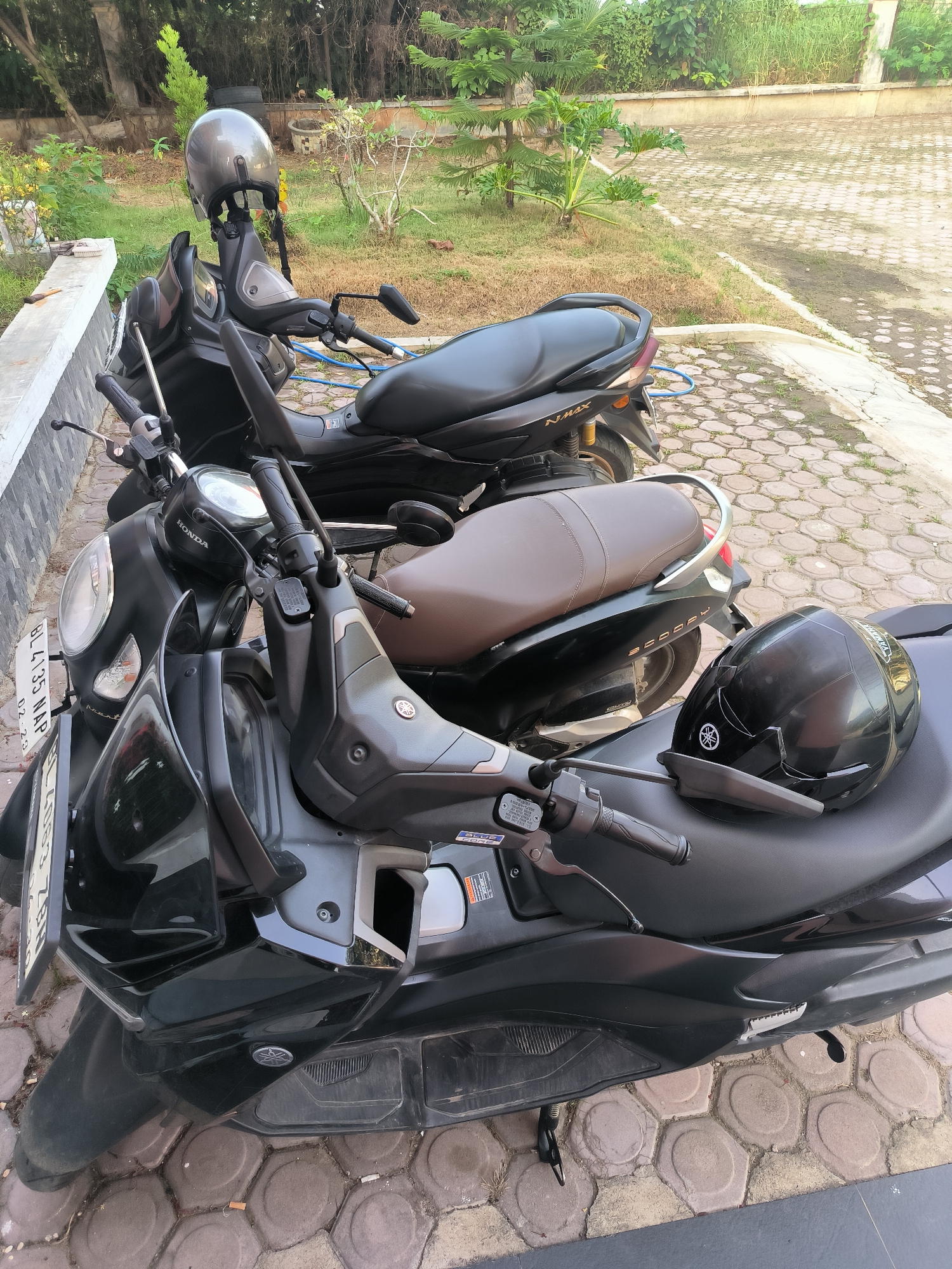 The image displays two black scooters parked on a paved surface composed of hexagonal tiles. The scooter in the foreground has a black helmet resting on its seat, and another grey helmet is perched on the handlebars of the scooter behind it. The scooters are parked outdoors, with lush green vegetation and trees visible in the background. A white concrete barrier runs along the left edge of the frame, separating the paved area from a grassy strip. A license plate that reads "BL 4135 NAP 02-23" is attached to the front of the foreground scooter. The scene appears to be in an urban environment, likely a parking area or walkway, within Lhokseumawe, Indonesia, given the style of the scooters and license plate.