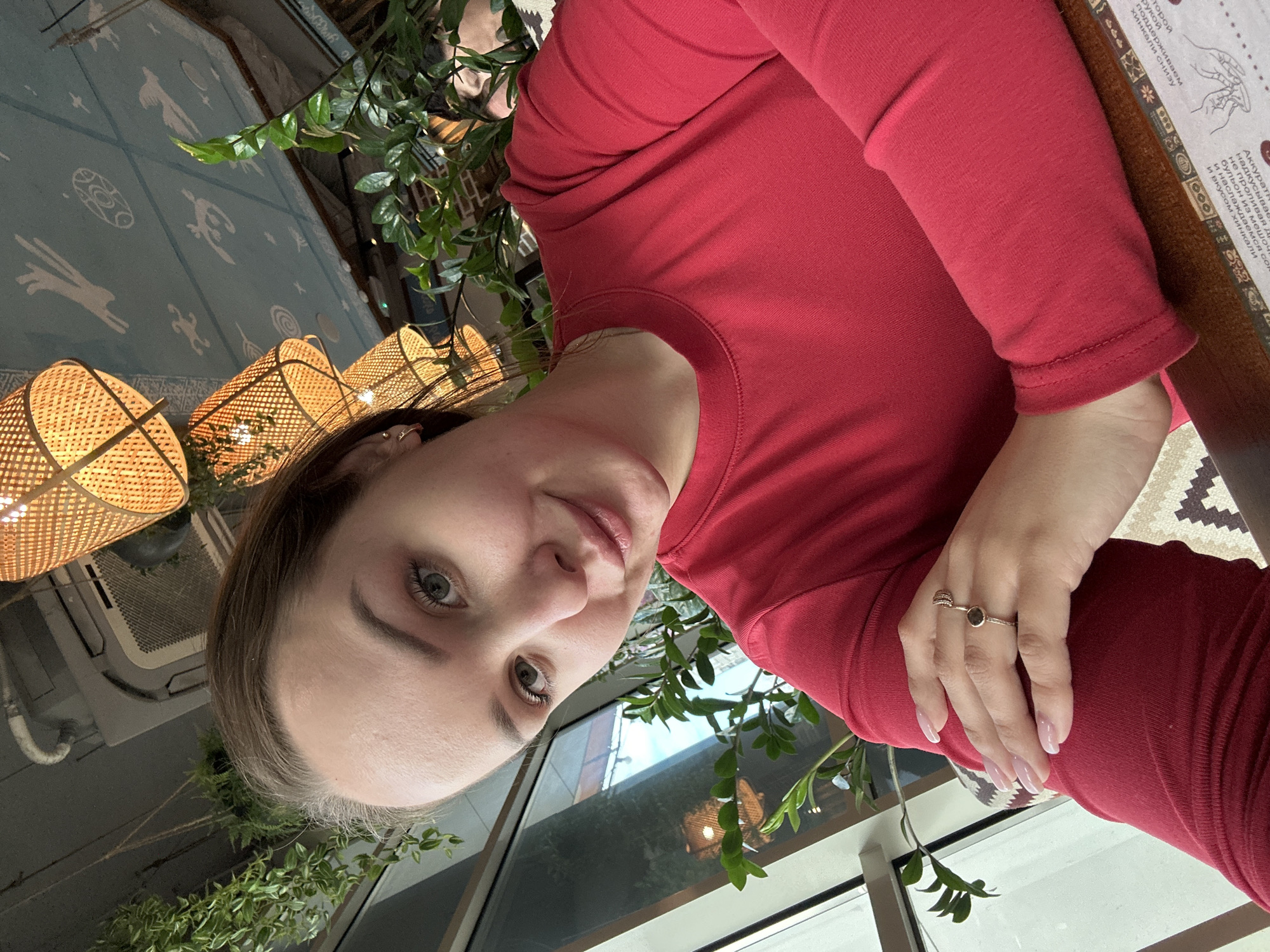 A fair-skinned woman with brown hair and wearing a long-sleeved red shirt is positioned in front of a window inside an establishment. Her left hand, adorned with two rings, is visible. The background features patterned walls, hanging wicker lamps, and decorative plants. Light from the window illuminates the scene. A printed menu is on the table. The location is Izhevsk, Russia.