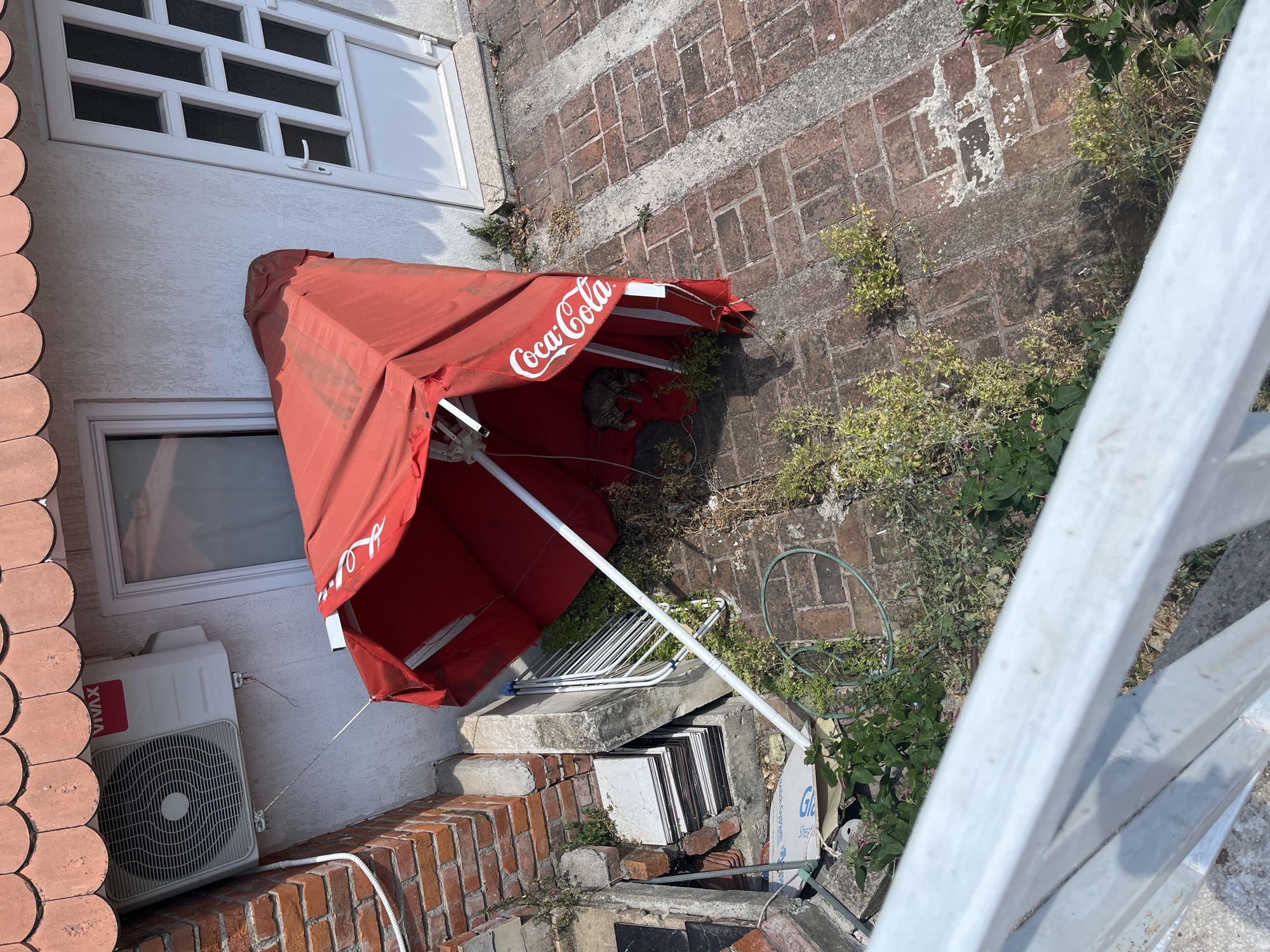 The image shows an outdoor scene with a red umbrella that has "Coca-Cola" written on it in white text. The umbrella is partially collapsed and resting on some steps. A cat is sleeping on the inside of the umbrella. To the left of the umbrella, there is a white air conditioning unit mounted on a wall. The wall is covered with light-colored textured plaster and has a window above the air conditioner. Below the air conditioner, there is a brick wall. The ground in the scene is paved with bricks, and there are plants growing between them. In the background, there is a white railing. The scene appears to be outdoors during the daytime, under sunny weather conditions. The location context is Budva, Montenegro.