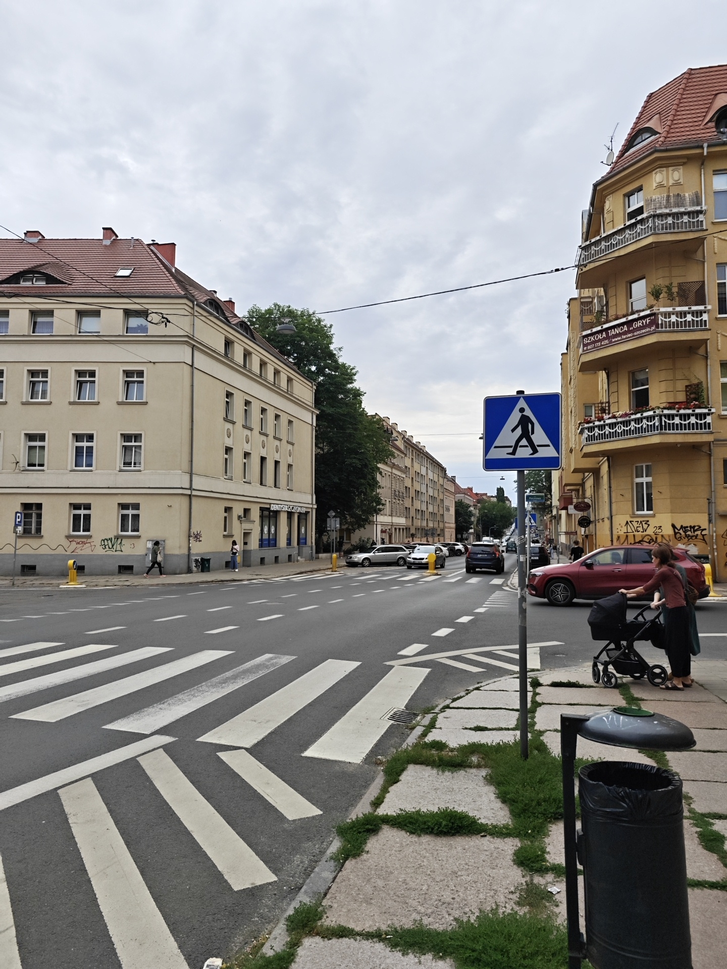 The image depicts a street scene in Szczecin, Poland, on an overcast day. In the foreground, a pedestrian crossing with white stripes is marked on the asphalt road. To the right of the crossing, a woman is pushing a stroller along a sidewalk. The sidewalk is made of concrete slabs interspersed with patches of grass. A dark trash can with an open lid stands near the woman.
Further down the street, cars are driving, and more pedestrians are visible. A blue and white pedestrian crossing sign is prominently displayed on a pole. On the right side of the street, a yellow building with arched windows and balconies stands tall, adorned with potted plants. A banner on this building reads "SZKOŁA TAŃCA „GRYF”" and includes a phone number and website. On the left side of the street, a cream-colored building with multiple windows and a red-tiled roof is visible. The overall atmosphere of the scene is ordinary and urban. The time of day appears to be daytime, given the ambient light, but the cloudy weather creates a muted lighting condition.