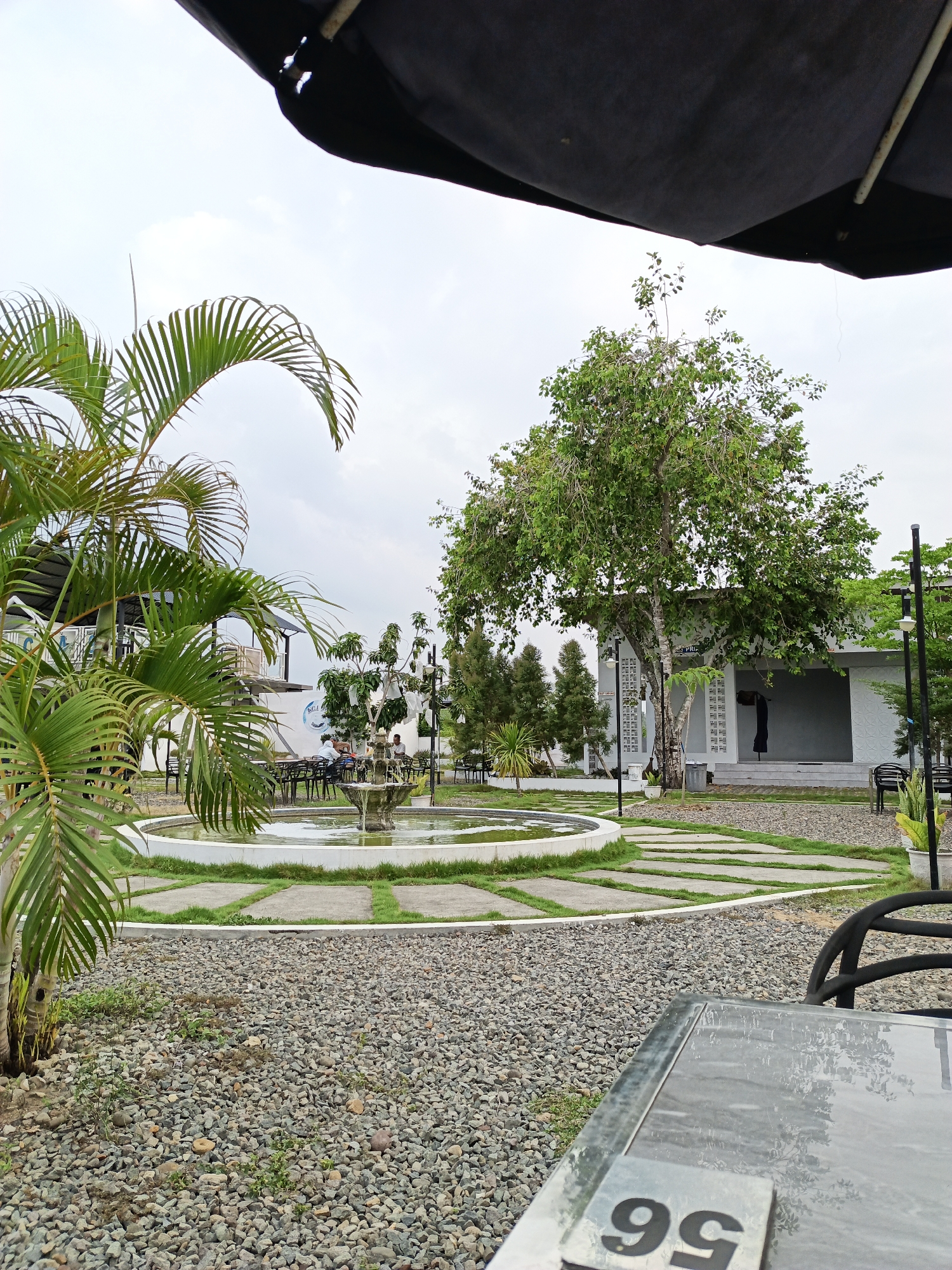 The scene depicts an outdoor area in Cotbadatunong, Indonesia. In the foreground, a table with the number "99" is visible. The ground is covered in gray gravel. To the left, there is a palm tree. In the middle ground, there is a circular fountain surrounded by square stepping stones with grass in between. Behind the fountain, there are trees and a white building with a gray roof. People are sitting at tables near the fountain. The sky is overcast. An unidentified black object is visible at the top of the image.