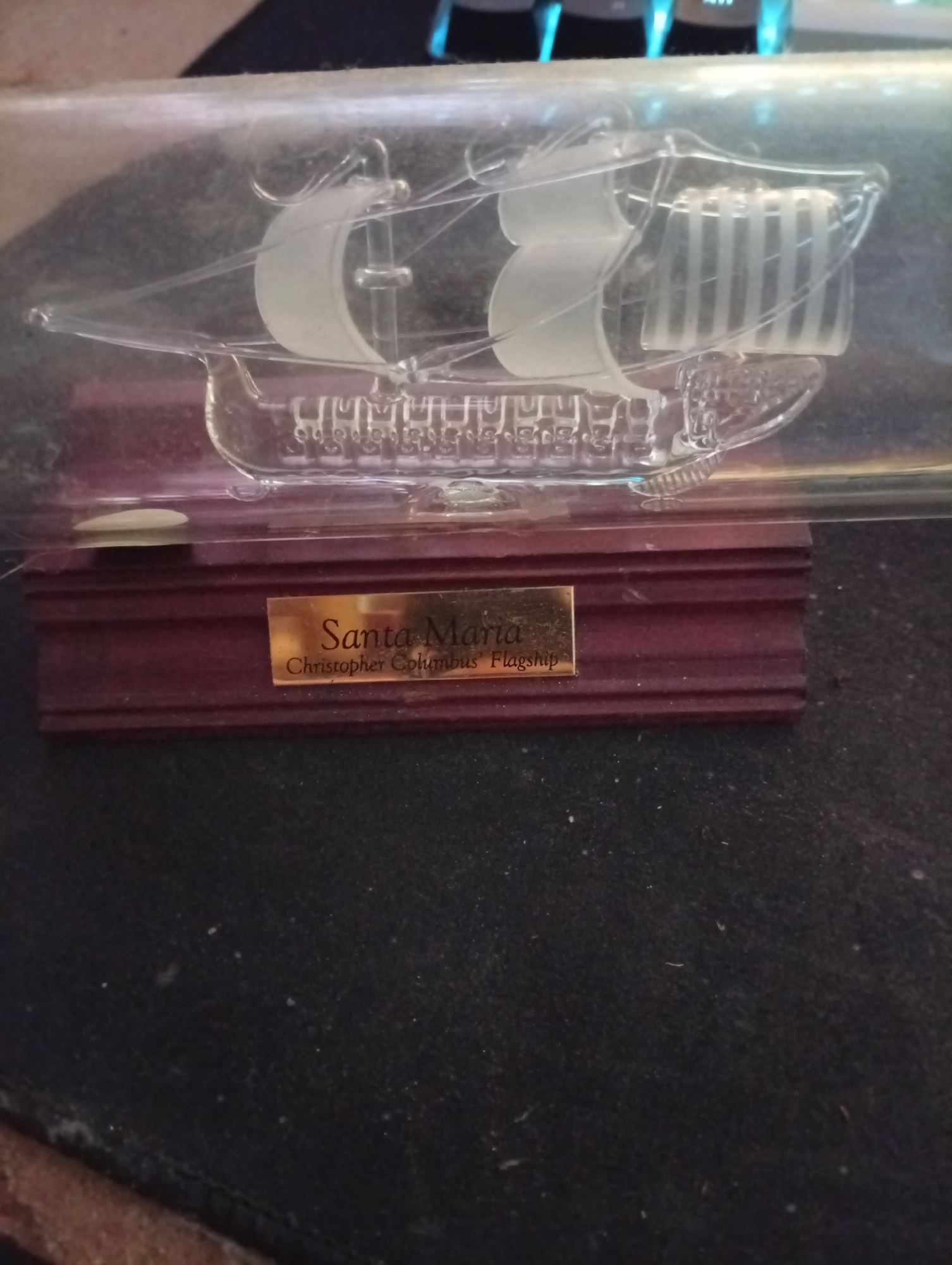This image shows a glass model of a sailing ship on a wooden base. The ship is transparent and has white sails. A gold-colored plaque on the base reads "Santa Maria, Christopher Columbus' Flagship". The object is placed on a dark, textured surface that appears to be fabric, possibly a desk or table, with some dust particles visible. The background is out of focus and shows what appears to be a keyboard with blue and white lights. There is no indication of the city of Wedel, Germany in the image.