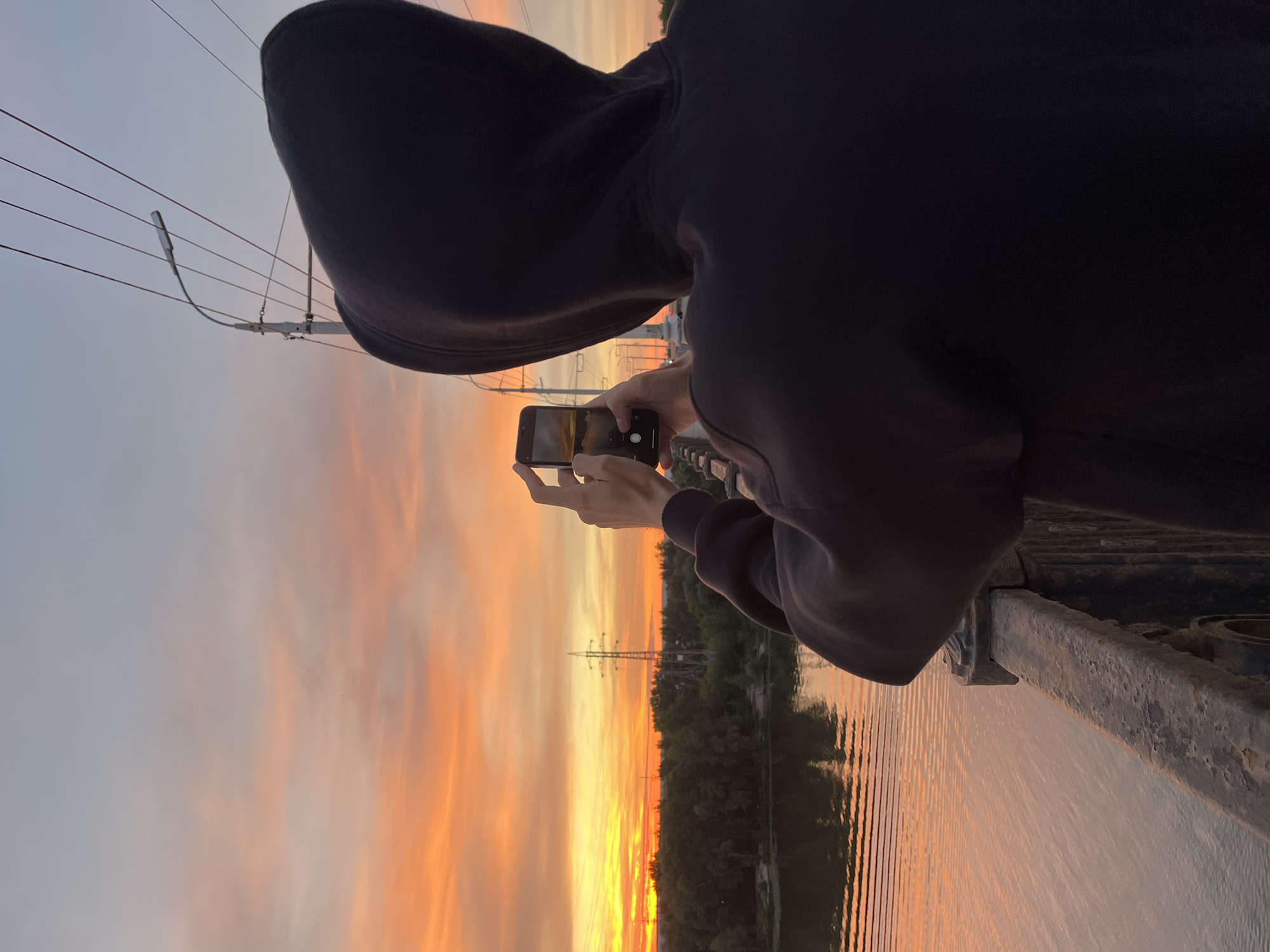 A person in a black hooded sweatshirt is holding up a smartphone to capture a photo of a sunset. The phone is oriented vertically, and the person's hands are visible as they operate the device. The sunset is vibrant, with hues of orange, yellow, and pink painting the sky. A body of water, likely a river, reflects the colors of the sunset. Trees and a power line tower are silhouetted against the colorful sky on the far bank of the river. The person appears to be standing on a bridge or elevated platform, as indicated by the metal railing on the right side of the image. The time of day is clearly sunset, and the weather appears clear. The overall mood is serene and appreciative of the natural beauty. No visible text is present in the image. The location is identified as Samara, Russia.