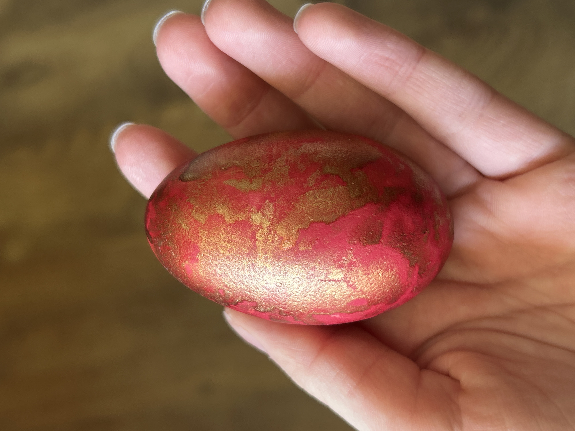 A hand holds a red and gold egg. The background is a blurry brown. The image does not show the city of Krumovo, Bulgaria.