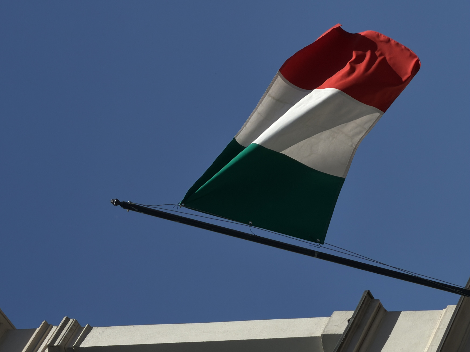 The image shows an Italian flag waving against a clear blue sky. The flag is attached to a dark pole, which is partially visible on the left side of the frame. The flag is composed of three vertical bands of color: green on the left, white in the middle, and red on the right. A portion of a pale, ornate building is visible on the far left of the image. There are no people or animals present in the scene. The image appears to be taken outdoors, in daylight. Based on the provided clues, the image was taken in Munich, Germany.