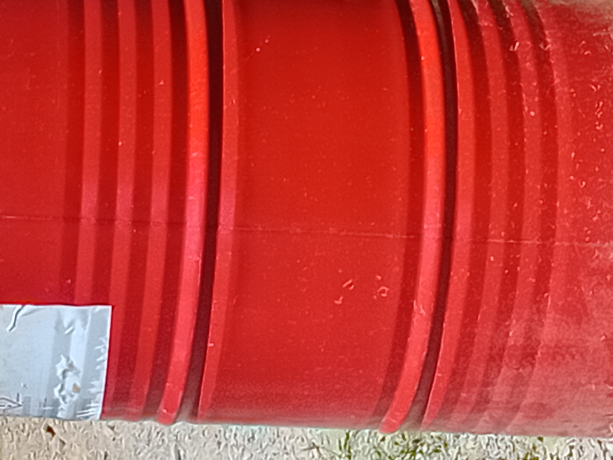 The image shows a close-up, vertical view of a red, ribbed barrel. A white label with some markings is visible on the upper left portion of the barrel. The background is blurred and appears to be a light-colored, textured surface, possibly gravel or sand, with some hints of green foliage on the left side. The scene is likely an outdoor setting, potentially in Verkhniachka, Ukraine, given the context of the prompt.