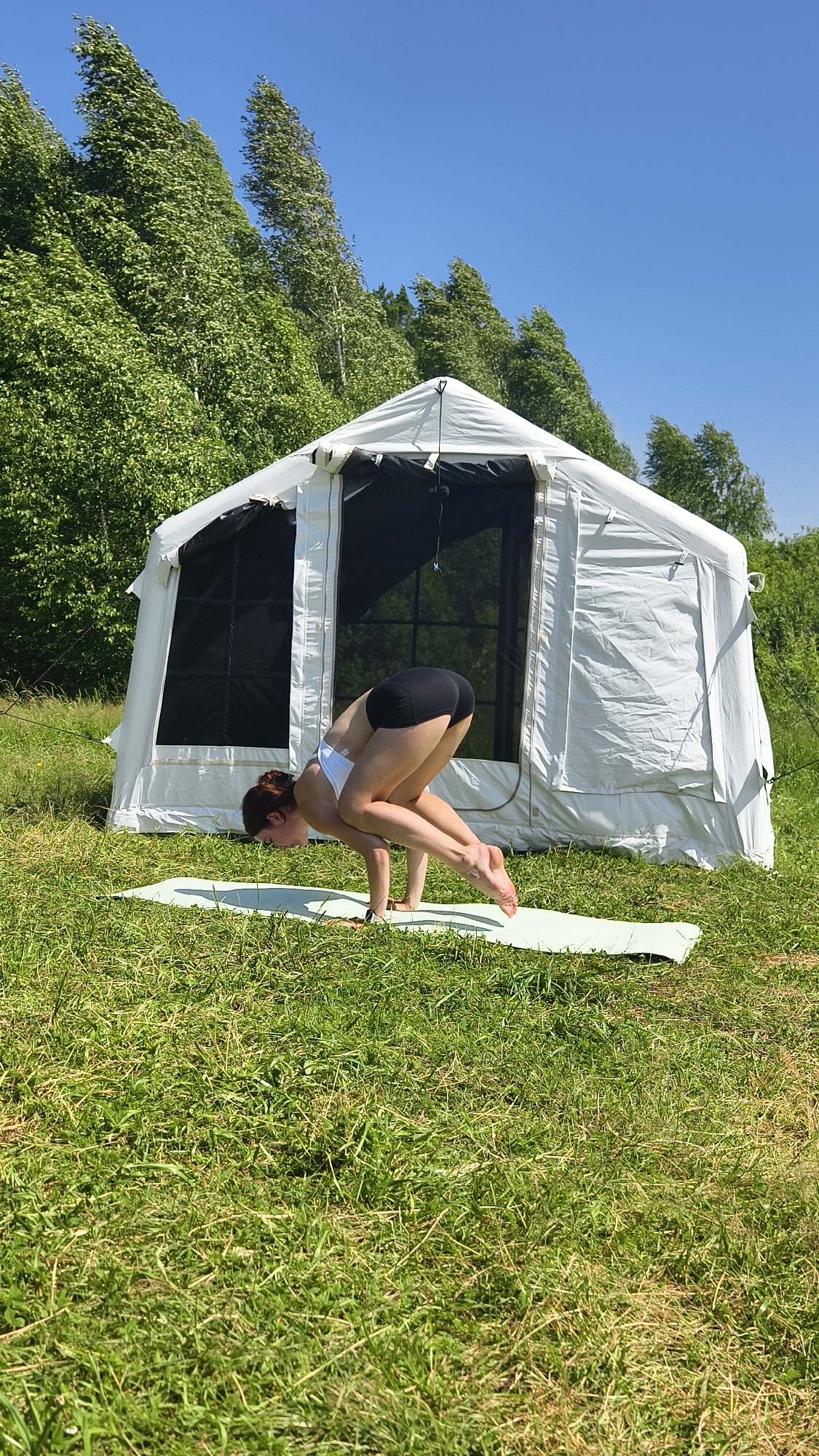 A woman is performing a yoga pose on a mat in front of a white tent in a grassy field. The woman is wearing a white sports bra and black shorts. She is balancing on her hands with her knees bent and tucked towards her chest. The tent is large and appears to be made of canvas. It has large windows with black mesh screens. The background consists of a lush green forest and a clear blue sky. The overall scene suggests an outdoor recreational activity in a natural setting. The location is identified as the city of Tyumen, Russia.