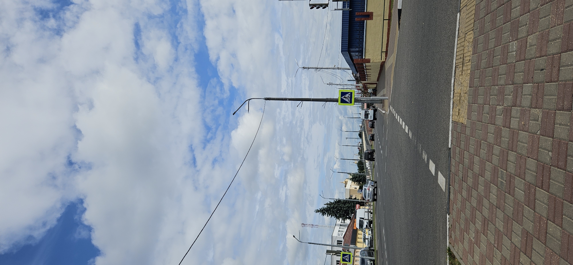 The image depicts a city street intersection in Vitsebsk, Belarus. The sky is partly cloudy with blue sky visible. On the right side of the road, there is a yellow building with a blue roof and a brick accent. A pedestrian crossing sign with a blue pedestrian symbol on a yellow background is visible near the intersection. Several cars and a white van are driving on the asphalt road. Trees and buildings are visible in the background. The foreground shows a brick-patterned sidewalk.