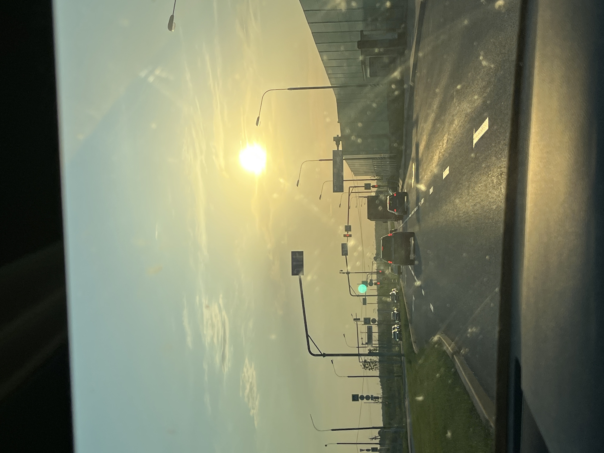 The image depicts a street scene, likely taken from inside a vehicle. The sun is visible in the sky, creating a bright and hazy atmosphere. A road stretches into the distance, with several cars and other vehicles visible on it. Streetlights and traffic signals line the road. The image captures the urban landscape and the movement of traffic under a bright sky.