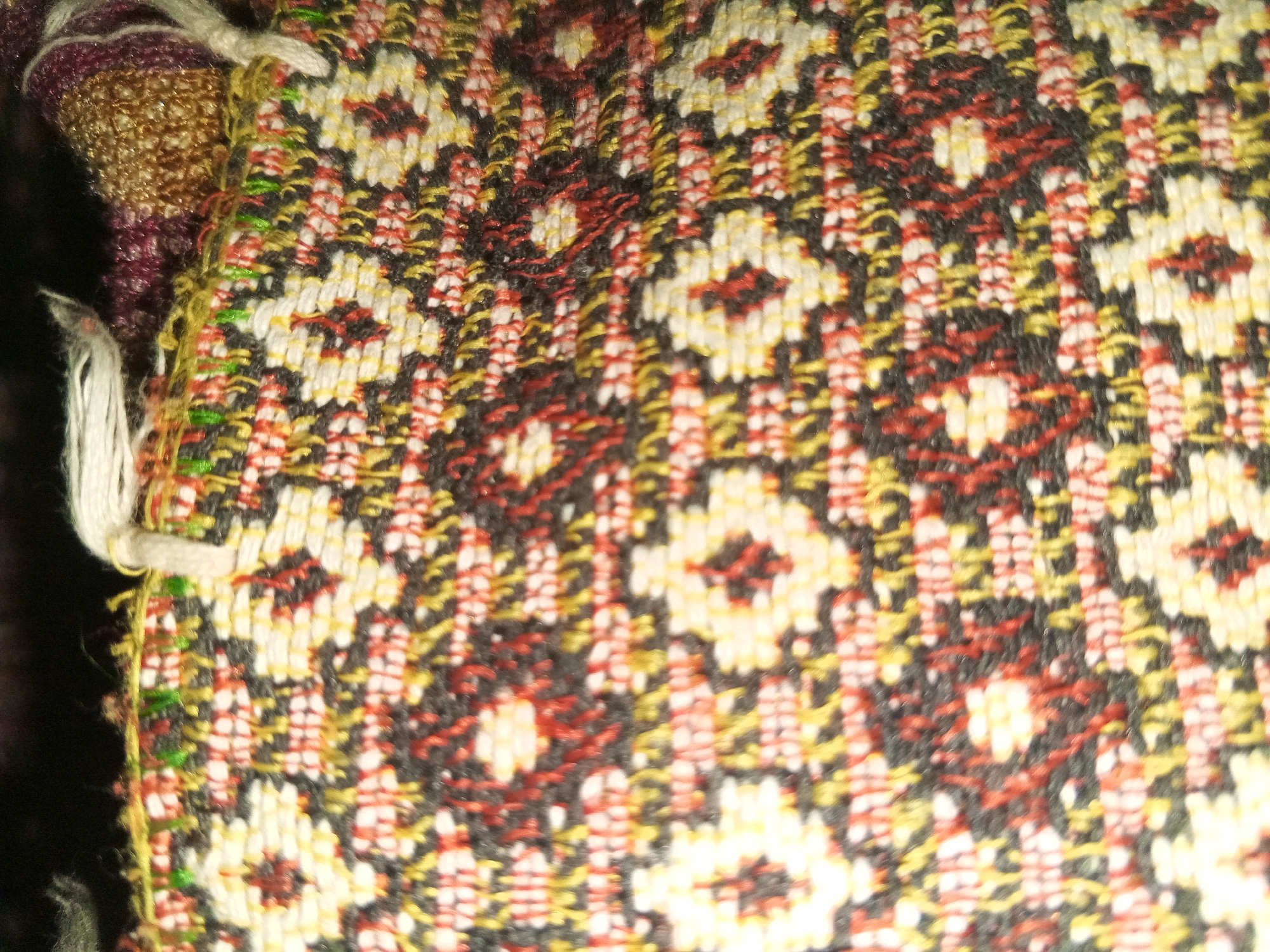 The image shows a close-up of a woven textile with a geometric floral pattern. The textile is composed of threads in various colors, including white, yellow, red, and black, creating a textured surface. The pattern features repeating motifs that resemble stylized flowers with multiple petals. The edges of the textile show fringed threads, indicating it might be a scarf, shawl, or decorative fabric. The background is dark and out of focus, suggesting the textile is the primary subject. There are no people or any elements that would indicate a specific location in Kanju, Pakistan.