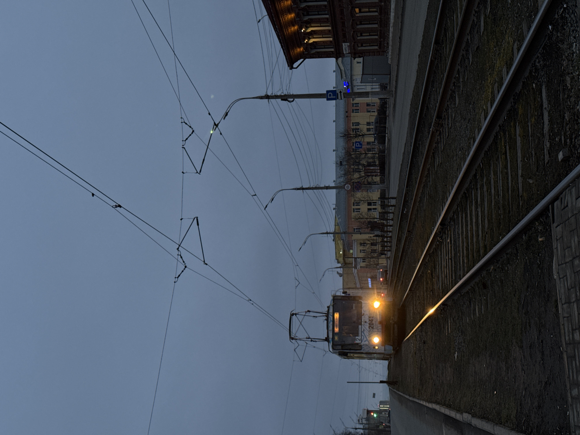 The image shows a streetcar approaching along a tram line in Izhevsk, Russia. The streetcar is identified by its number, 2041. The surrounding environment consists of a building with parking signs, suggesting an urban area. Overlapping the streetcar and tracks are several power lines. The sky is overcast.