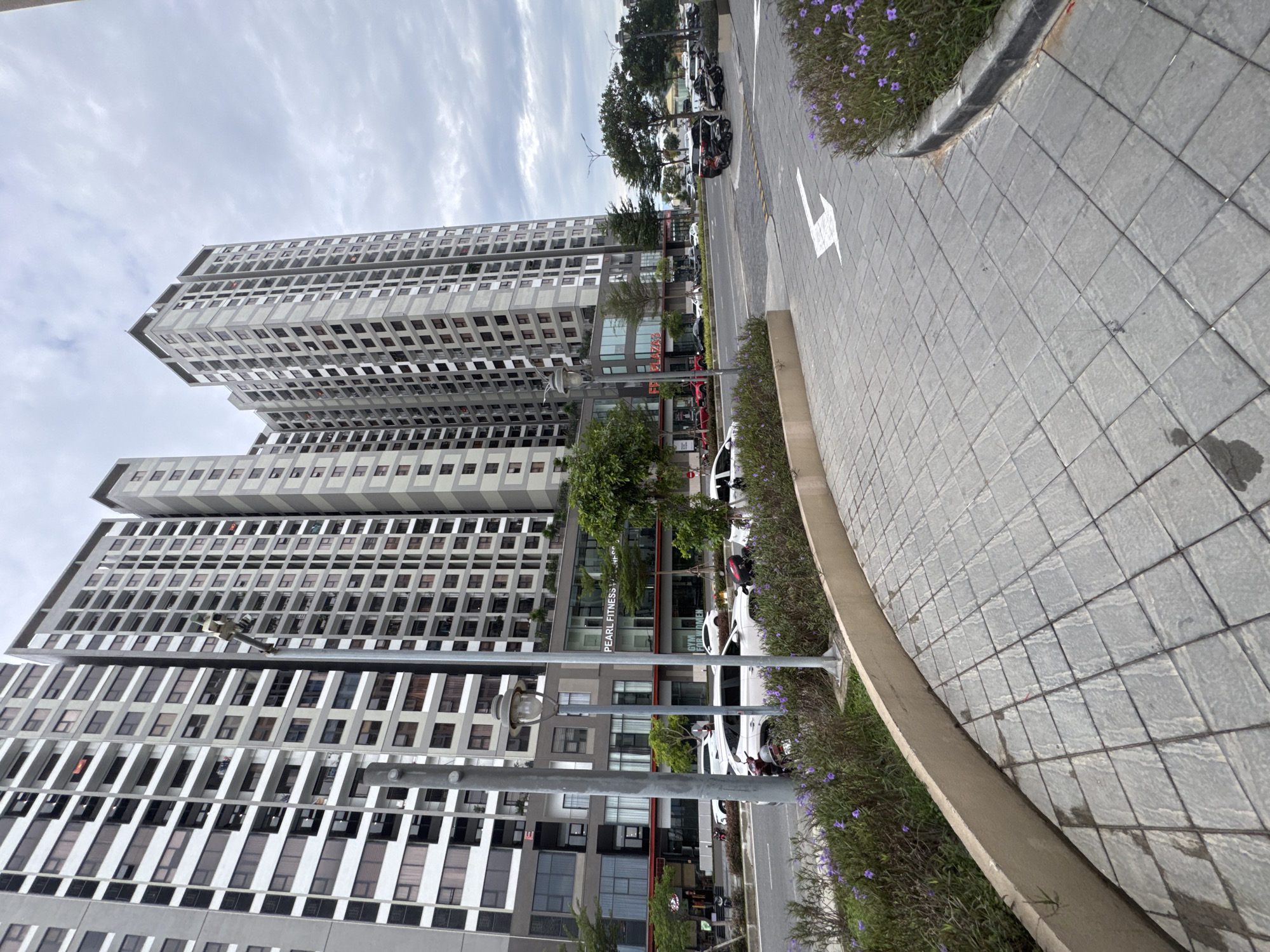 The image shows a street scene in Da Nang, Vietnam, with several modern, high-rise apartment buildings dominating the background. In the foreground, a paved walkway curves to the right, bordered by low-lying green bushes with small purple flowers. The walkway is made of square gray pavers laid in a diamond pattern. A concrete curb separates the walkway from the street. Across the street, several cars and motorcycles are parked or driving. The buildings are tall, with numerous windows and balconies. Some of the buildings have commercial spaces on the ground floor, including a sign that reads "PEARL FITNESS". The sky is overcast with clouds. A street lamp is visible in the middle left of the image, with metal poles supporting it. The overall impression is one of urban development and a busy street.