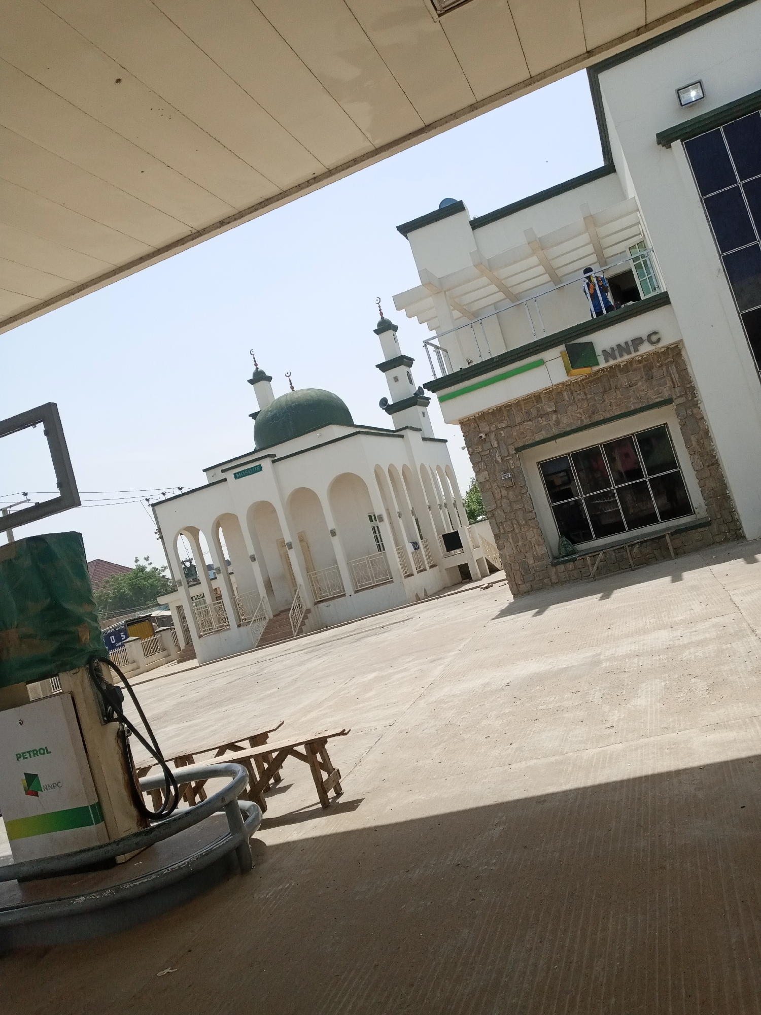 The image shows an outdoor scene with a petrol station and a mosque. In the foreground, a white petrol pump with "PETROL" and "NNPC" branding is visible. A green tarp is draped over a portion of the pump. To the right of the pump, there are two wooden benches.
In the mid-ground, a white mosque with a green dome and minarets stands prominently. The mosque features arched entrances and white railings. Adjacent to the mosque is a building with "NNPC" signage and a stone-clad section with a large, multi-pane window. A person in a blue and yellow uniform is standing on a balcony of the NNPC building.
The ground is paved with concrete. The sky is clear and bright, suggesting a daytime setting with sunny weather. The overall scene appears to be a commercial area where a petrol station and a mosque are located in close proximity. There is also a hint of other buildings and trees in the background.
Visible text includes "PETROL" and "NNPC" on the petrol pump, and "NNPC" on the building. The word "Mosque" is also visible above the entrance of the mosque.