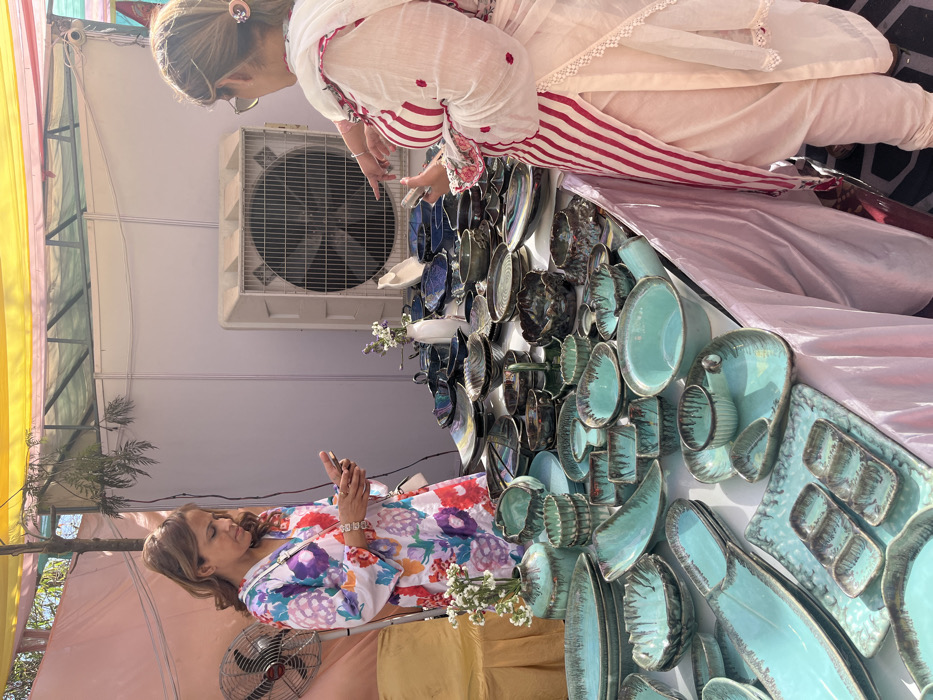 The image shows two women standing at a table displaying a variety of ceramic dishes and bowls at a market in Sector 17, Chandigarh, India. One woman, wearing a white dress with floral patterns, holds a phone in her left hand and appears to be looking at it. The other woman, wearing a white dress with red stripes, points her right hand at the table. The table is covered with a white cloth and lined with an array of teal-colored ceramic dishes and bowls with dark streaks. A large industrial fan is visible in the background. The setting is outdoors with a canopy overhead.