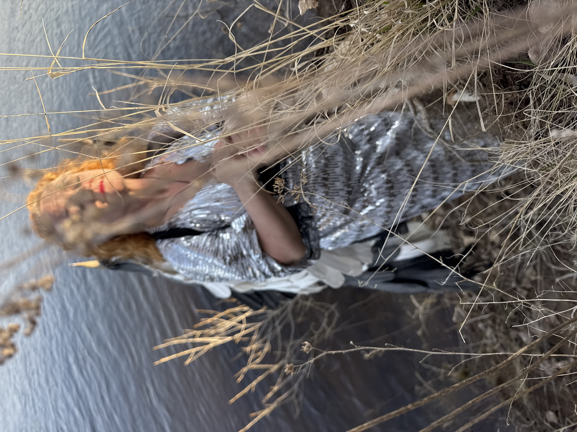 A woman with red hair and red lips is wearing a silver sequined dress and laying on the ground next to water. Dried reeds and grasses surround her. The location is likely near a body of water, and the season appears to be late autumn or early winter. The photo's angle gives the impression the woman is reclining. There are white garments beneath her, and a black bag is nearby. There is no confirmation the location is Noviy, Russia.