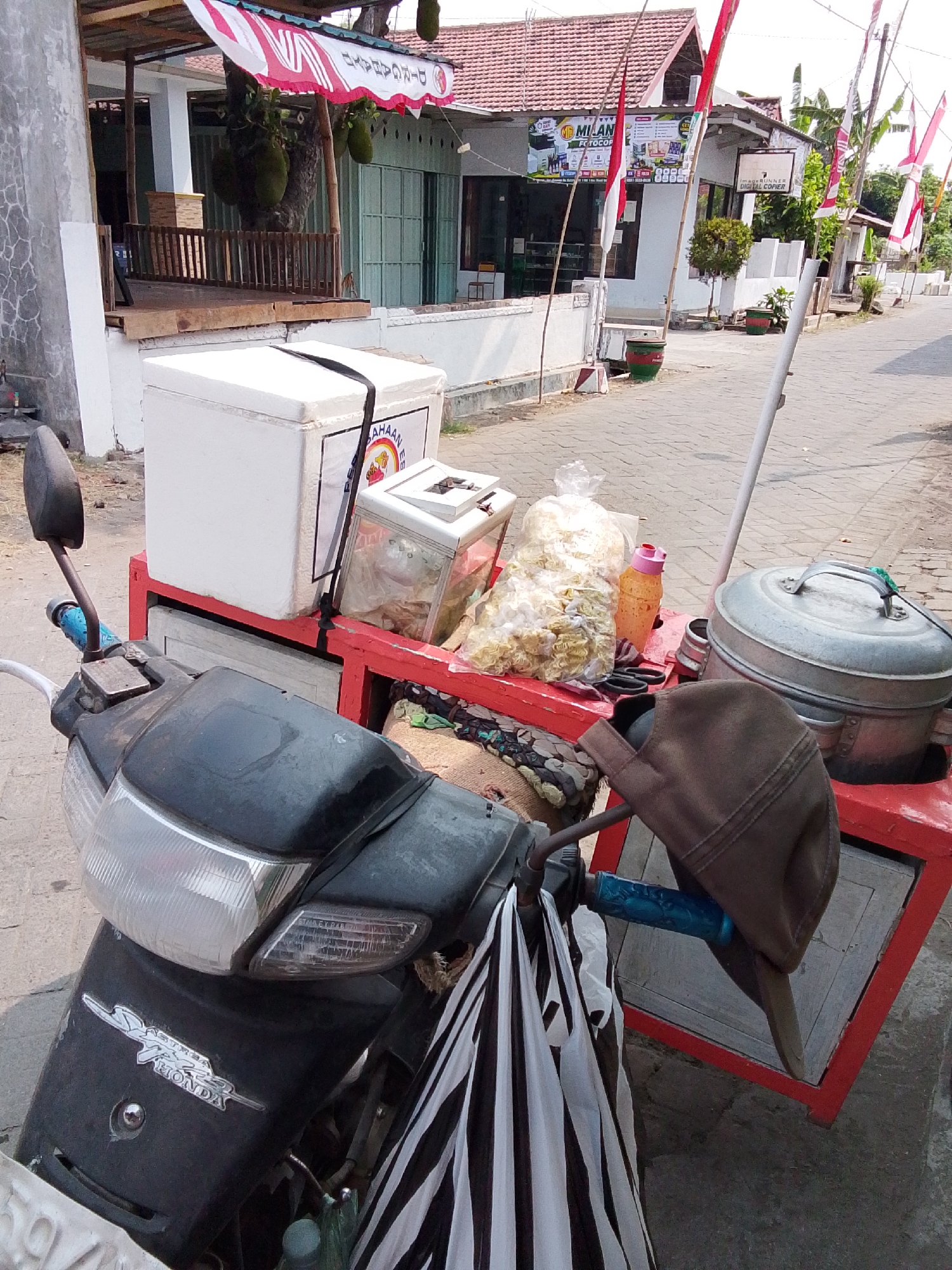 In Bulurejo, Indonesia, a parked motorcycle, a black Honda Astrea, is positioned in the foreground, facing left. A red cart is attached to the motorcycle, holding a white cooler, a smaller display case with food items, a large bag of snacks, a bottle of orange liquid, and a metal steaming pot. A brown cap rests on the cart, and a striped bag hangs from the motorcycle's handlebars. In the background, a building with a red tile roof and an awning displays a banner with Indonesian text and logos. Several Indonesian flags are visible along the street, indicating a festive or patriotic occasion. The street is paved with grey bricks, and the environment appears to be a typical street scene in a small Indonesian town.