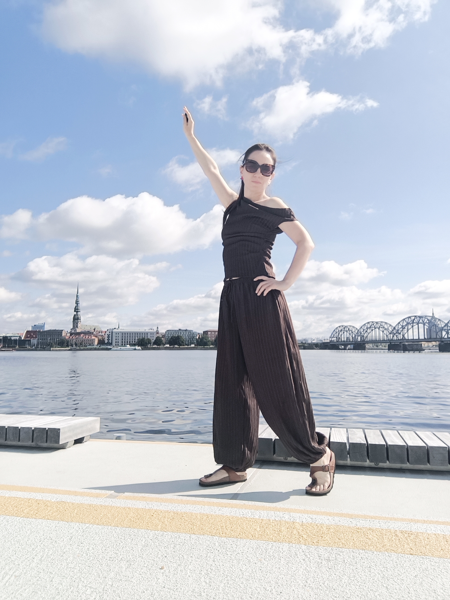 A woman stands on a paved walkway by the water in Riga, Latvia, with her right arm raised. She is wearing a dark brown off-the-shoulder top and loose-fitting pleated pants, along with brown sandals. She has on sunglasses. The walkway is bordered by yellow lines and has a wooden bench to her left. Behind her, a body of water is visible, with the Riga city skyline in the distance, featuring distinctive architecture including a tall spire. A large arched railway bridge is also visible across the water on the right side of the frame. The sky above is blue with scattered white clouds.