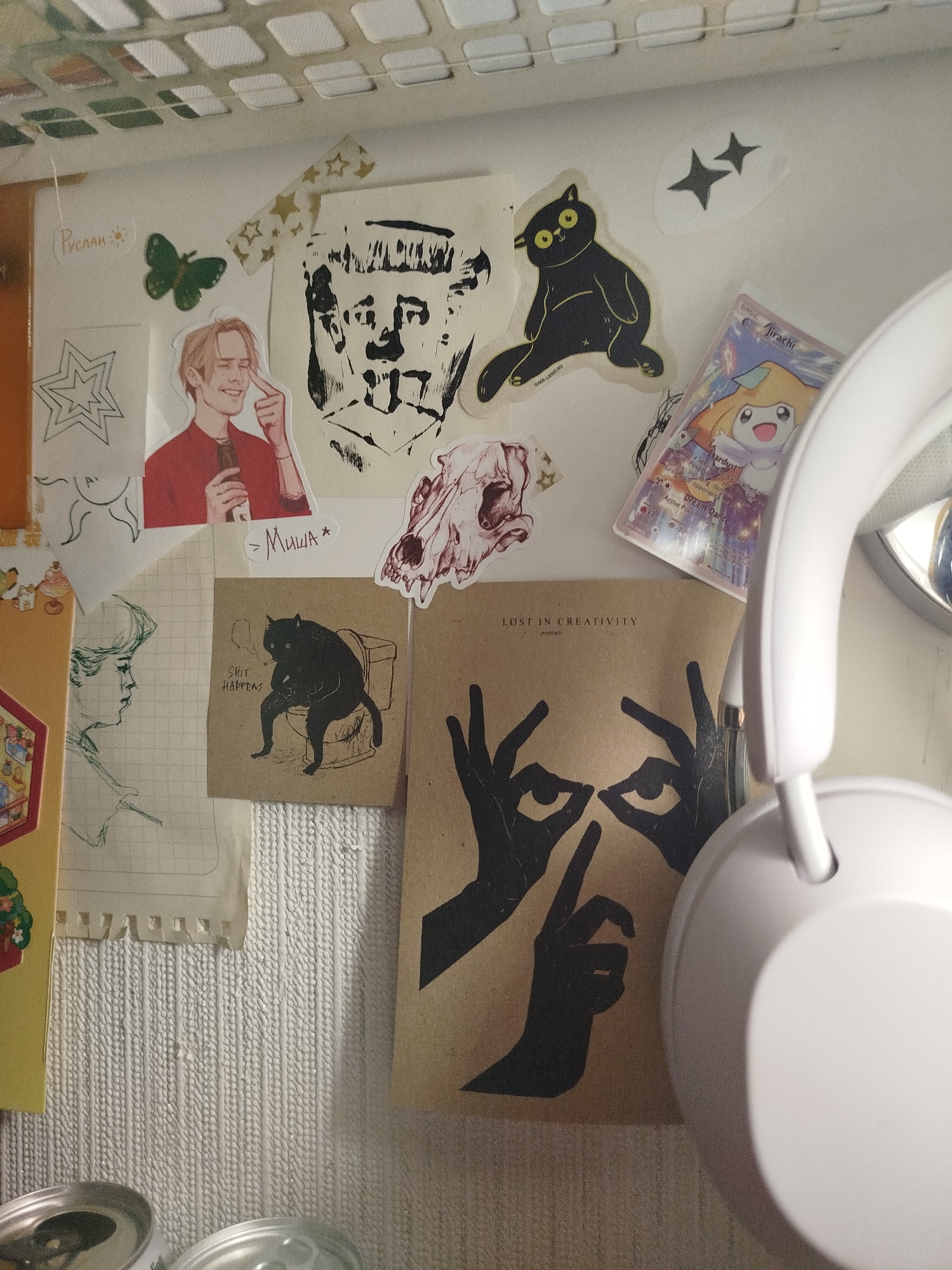 The image shows a wall or surface covered in various stickers and drawings, partially obscured by a white pair of headphones on the right side. Stickers include drawings of cats, a portrait of a man, a human skull, and a Pokémon card featuring Jirachi. There are also drawings of stars and a butterfly. Text on some of the stickers reads "Руслан", "Миша", "SHIT HAPPENS", and "LOST IN CREATIVITY". Two cans are visible at the bottom of the image.