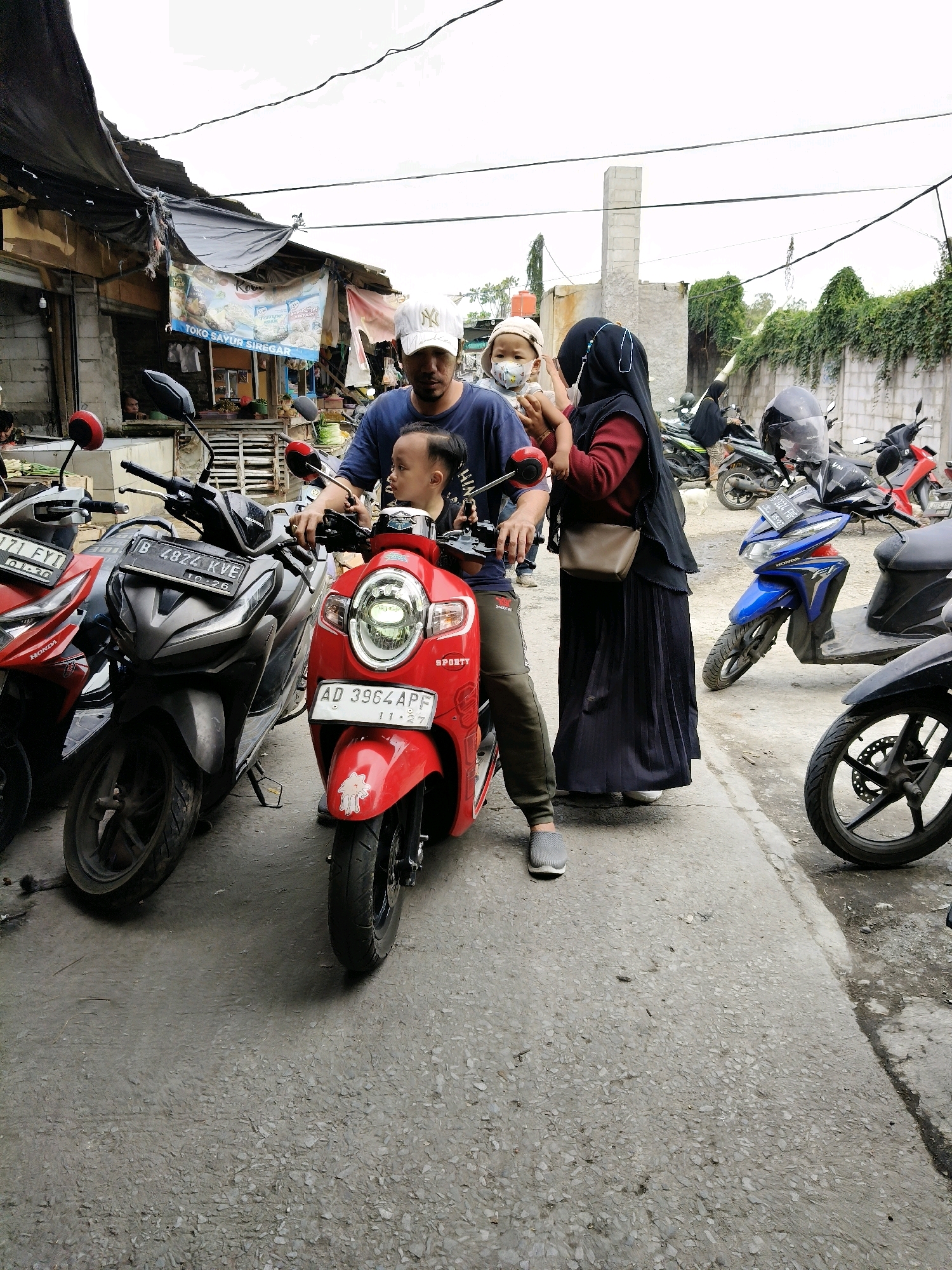 In Setiamekar, Indonesia, a family stands near several parked motorcycles on a street. A man in a blue t-shirt and a white baseball cap is seated on a red scooter, holding a young child on his lap. The child is wearing a light-colored cap and a face mask. A woman in a black hijab and a dark, pleated skirt stands beside the man, holding the child's hand. The background shows a row of market stalls with awnings and a brick wall with overgrown vines. Other motorcycles are parked on the street, including a dark grey scooter in the foreground. The ground is paved with asphalt.