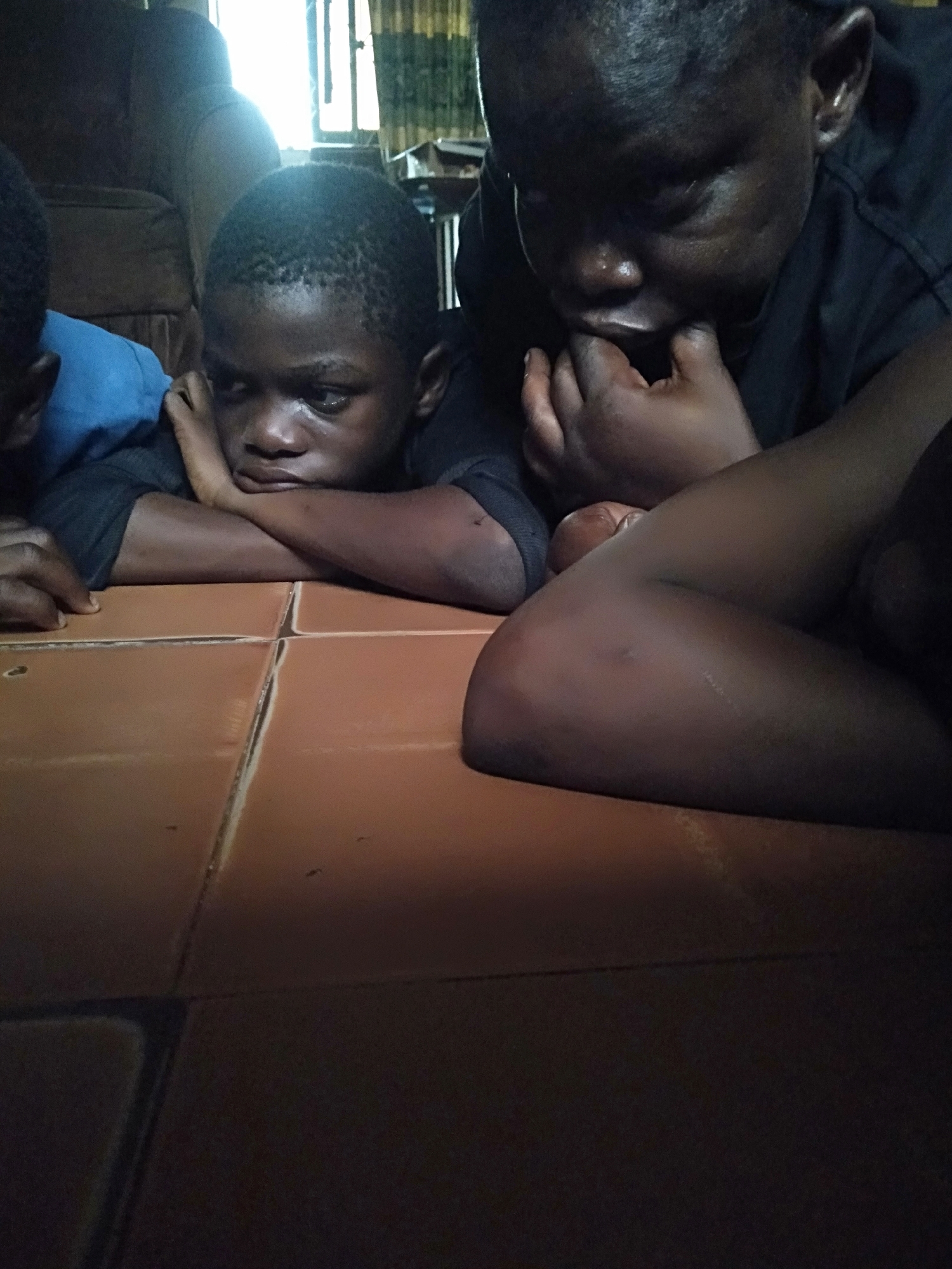 Three children are lying on a tiled floor. One child is in the foreground, resting his head on his crossed arms, with a melancholic expression. Another child is next to him, with his fingers in his mouth, seemingly in deep thought. A third child is partially visible on the left edge of the frame. The setting appears to be indoors, possibly in a residential area within Shangisha, Nigeria. There is a sofa partially visible in the background, along with some window coverings.