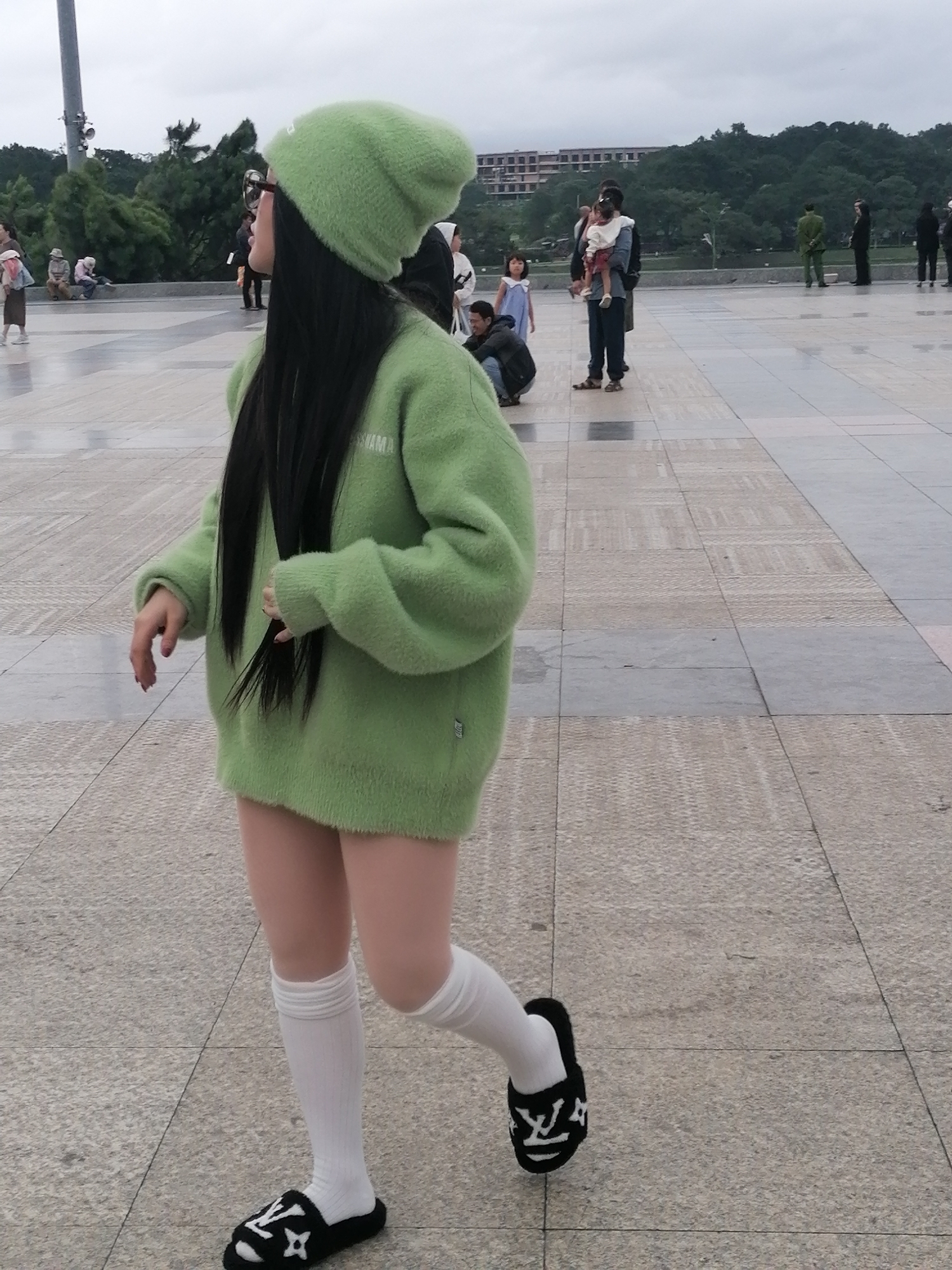 A young woman with long, dark hair is seen from the side, walking in what appears to be a public plaza in Da Lat, Vietnam. She is wearing a fuzzy, lime-green beanie and a matching oversized green sweater that extends to her upper thighs. Below the sweater, she has on white knee-high socks and black slide sandals with a white logo resembling a stylized "LV". She is looking to her left and her left arm is bent and held across her body, with her hand resting on her sweater. Her right arm is extended slightly behind her.
The plaza is paved with large, square tiles. In the background, there are other people, some sitting on a ledge, others standing or walking. A family is visible with a man holding a small child, and another child standing nearby. Beyond the plaza, there are trees and buildings. The sky is overcast. The overall scene suggests a casual outing in a city environment.