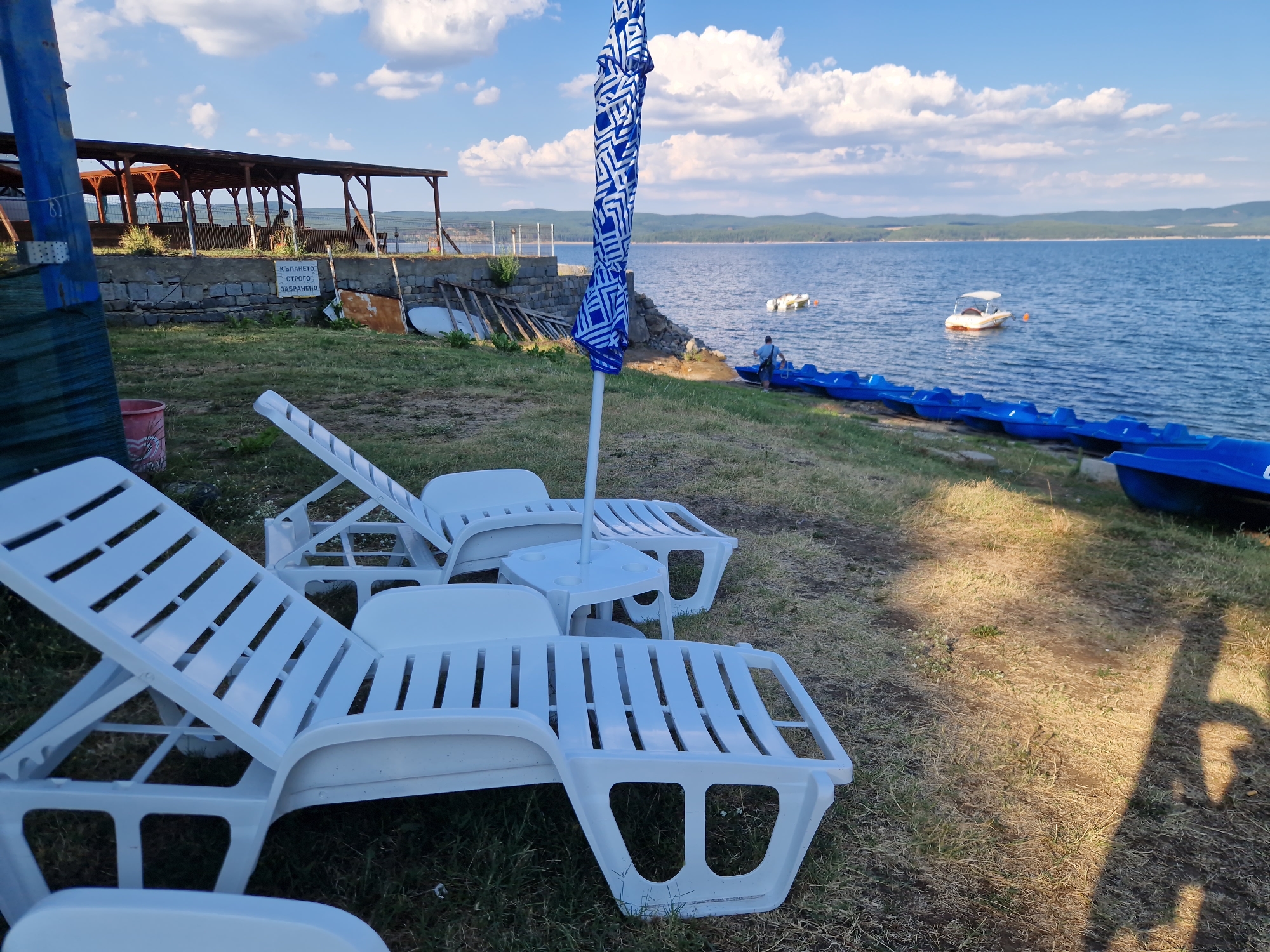 The image depicts a serene lakeside scene at the Iskar Dam, near Dolni Pasarel, Bulgaria. In the foreground, two white plastic lounge chairs are positioned on dry grass, with a small white side table and a blue and white patterned umbrella between them. To the right, a line of blue paddle boats is docked along the grassy shore of the tranquil blue water. The lake is dotted with a few small boats in the distance, and rolling, tree-covered hills form the backdrop under a sky with scattered white clouds. Behind the lounge chairs, a stone wall separates the grassy area from a wooden structure that appears to be a covered seating area or restaurant. A sign on the wall, written in Cyrillic, reads "КЪПАНЕТО СТРОГО ЗАБРАНЕНО" which translates to "SWIMMING STRICTLY FORBIDDEN." A person in a blue shirt can be seen near the paddle boats on the right side of the image, interacting with one of the boats. The overall impression is one of a quiet, undeveloped recreational area by the water.