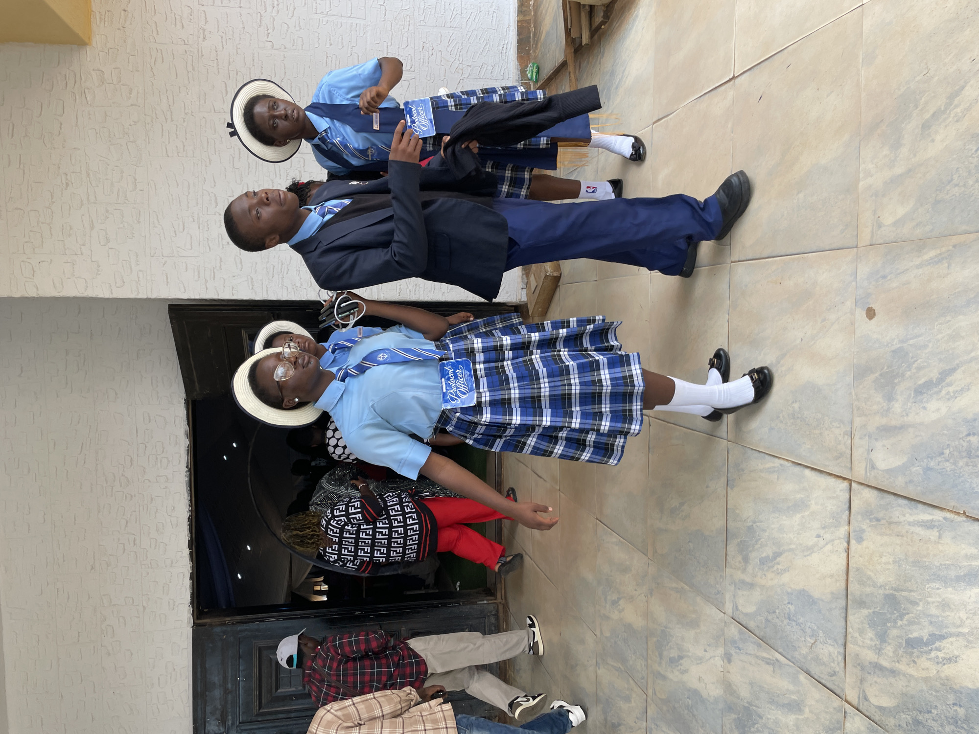 In Bar Gada, Nigeria, outside a building with a textured white wall and a dark doorway, three individuals in school uniforms are visible. Two females and one male, all appearing to be teenagers, are the primary subjects. The two females are wearing light blue shirts, blue and white plaid skirts, white socks, and black shoes. Both are also wearing white hats with black bands. One female is wearing a blue vest with a "Protocol Officer" badge. The male is in a dark blue suit jacket, a light blue shirt, dark blue pants, and black shoes. He is also wearing a dark vest underneath his jacket. The individuals are walking on a tiled floor that appears to be a light beige color with grey marbling. In the background, through an open doorway, several other people are visible inside a dimly lit space, some of whom appear to be wearing similar uniforms or formal attire. To the right of the frame, some wooden planks are stacked. The lighting suggests it is daytime, with the sun casting some shadows.