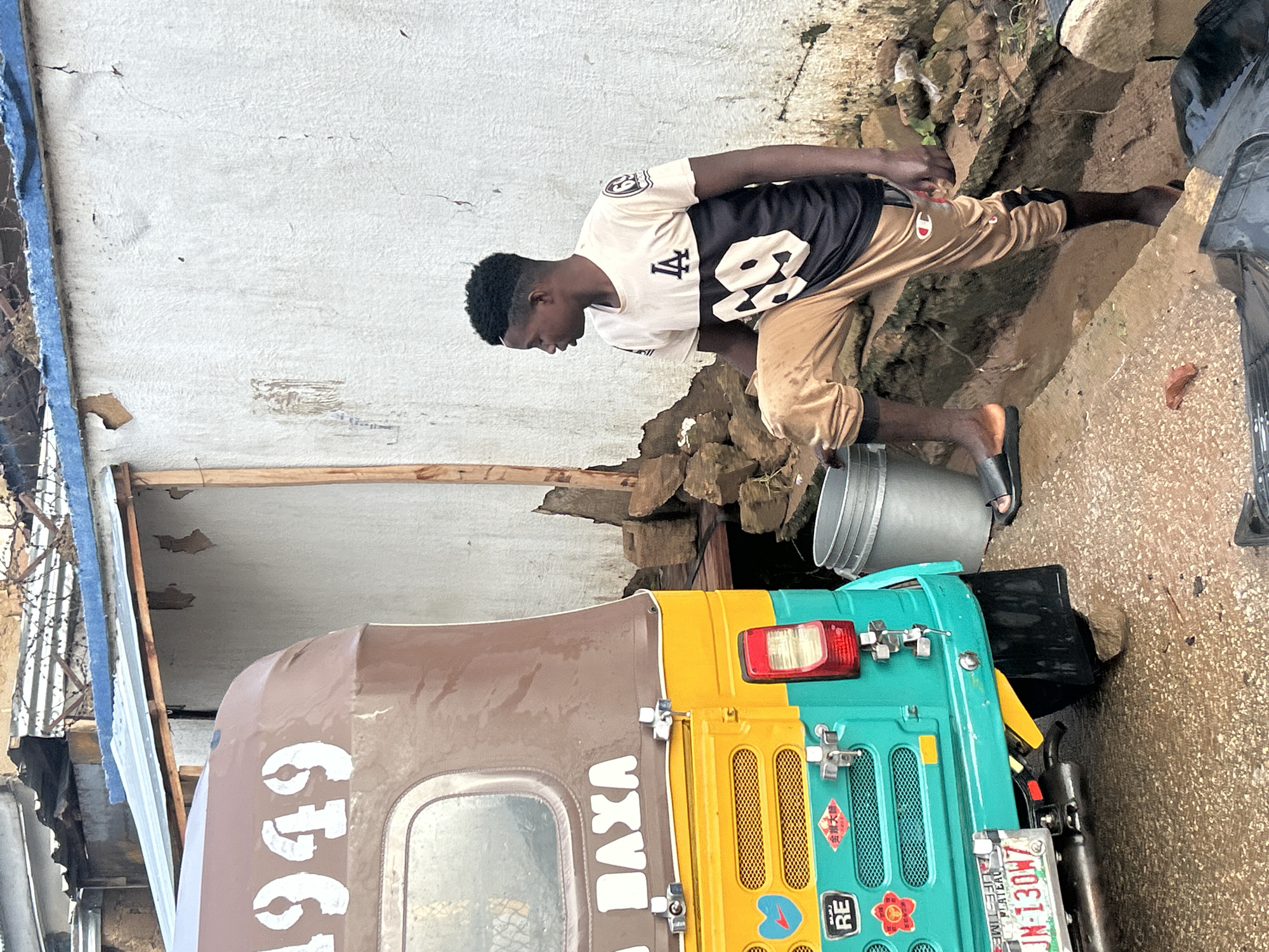 A young Black male stands to the right of a yellow and turquoise auto-rickshaw, facing left and looking downwards. He is wearing a cream-colored t-shirt with a black design featuring "LA" and the number "69," and tan cargo pants. His right leg is bent and resting on a raised dirt embankment, and his left foot is in a black sandal partially submerged in muddy water. To his left, a silver metal bucket is positioned on the embankment. The auto-rickshaw is parked on a wet, pebbled surface. The auto-rickshaw has the number "1949" and "DAYA" stenciled on its upper portion in white. A license plate that reads "JN-130W7" is visible on the rear of the vehicle. The background shows a dilapidated wall with peeling paint and a corrugated metal roof with barbed wire on top. The scene appears to be in Jos, Nigeria, characterized by the urban decay and the presence of the auto-rickshaw, a common mode of transport in the region.