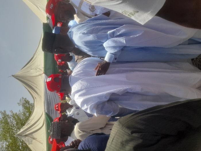 The scene is a political rally in Gusau, Nigeria, located at coordinates 12.156 latitude and 6.71 longitude. The event is being held under large, white, tent-like structures. The men are wearing white and blue garments with some wearing red caps that say "PDP" on the front. There are a number of men wearing white caps with a white fabric over them. The men are standing close to each other with arms around each other, likely showing solidarity and support for a candidate or party. The men appear to be engaged in a conversation and some are smiling while others are looking around. It appears to be a bright, sunny day. The ground is dry and appears to be dirt. A few people are standing around in the background. The scene is focused on the group of men in the center. The men are all of African descent.