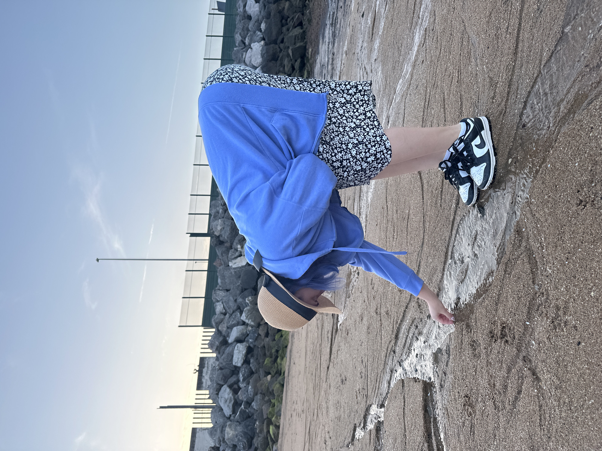 A person wearing a blue hooded sweatshirt, a short black and white floral dress, and black and white Nike Dunk sneakers stands on a sandy beach with water running over the sand. The person is bending over, reaching out with their right hand to touch the water. They are wearing a straw hat with a black band. Behind them is a seawall made of large rocks. The sky is light blue. The location appears to be a beach near a seawall in Gijón/Xixón, Spain.